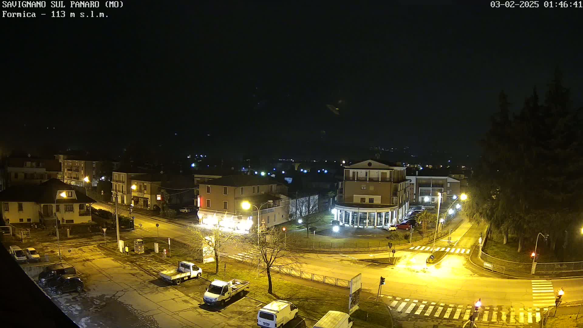 An aerial nighttime view of a town square with parked cars, streetlights illuminating the area, and a dark sky.