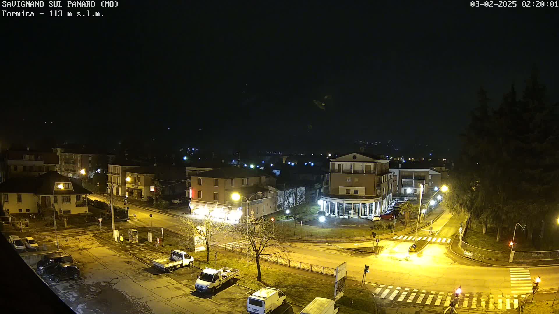 A nighttime aerial view of a town square with several buildings, parked cars, and streetlights illuminating the wet roads under a clear, dark sky.