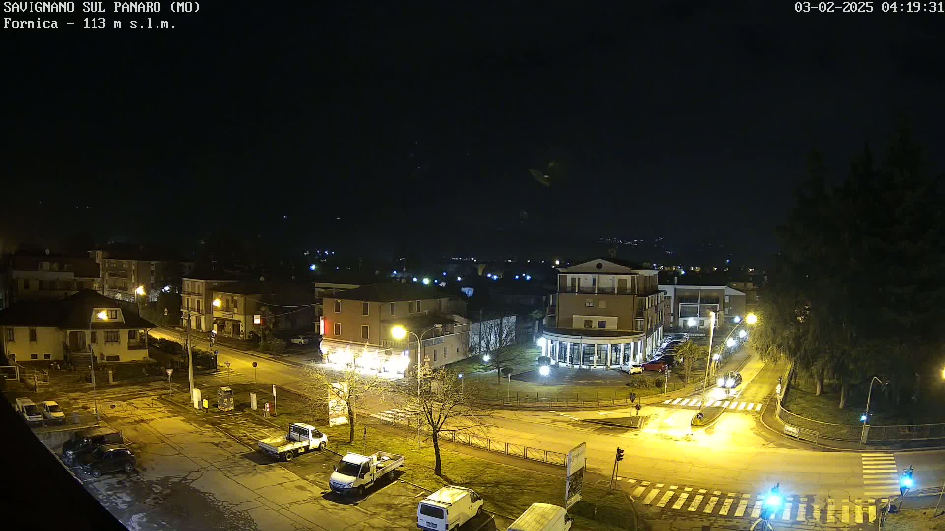 A nighttime aerial view of a town square with parked cars and buildings illuminated by streetlights under a clear, dark sky.