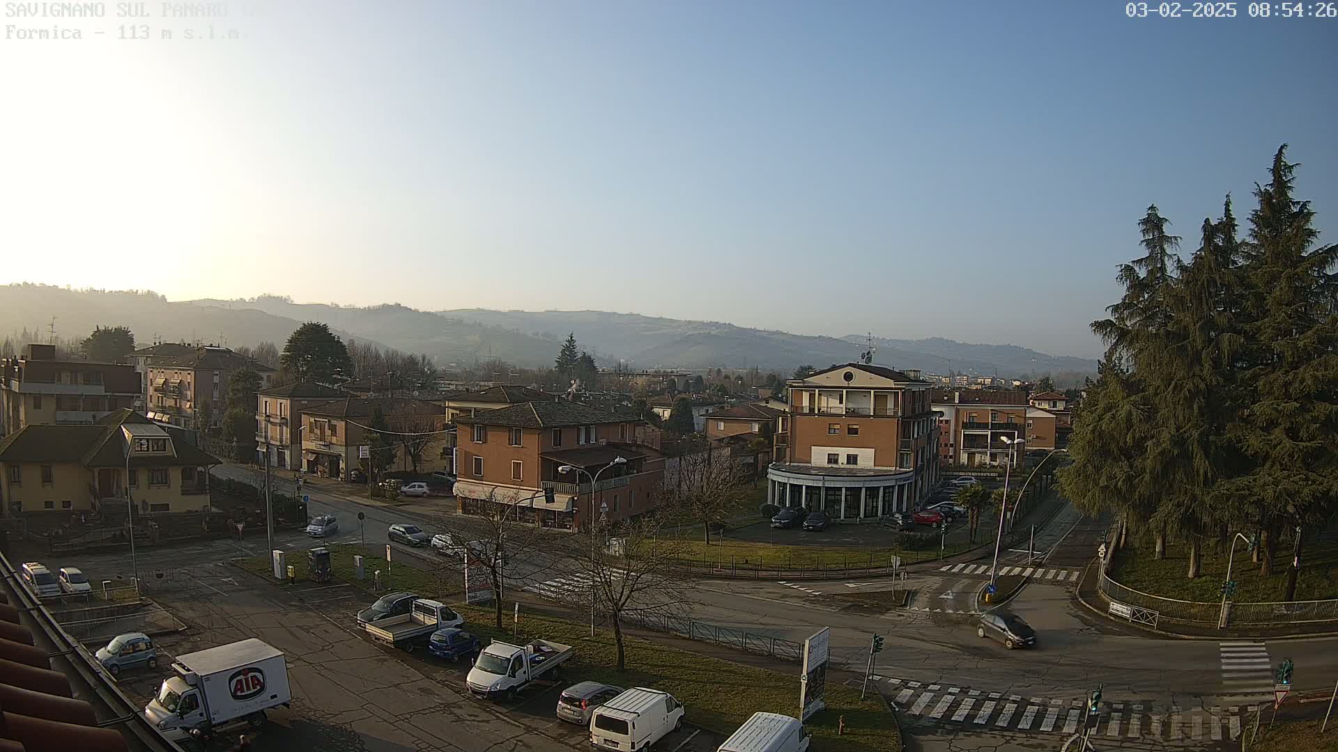 A hazy, sunny day overlooks a town square with several buildings, cars, and evergreen trees, all nestled against a backdrop of rolling hills.