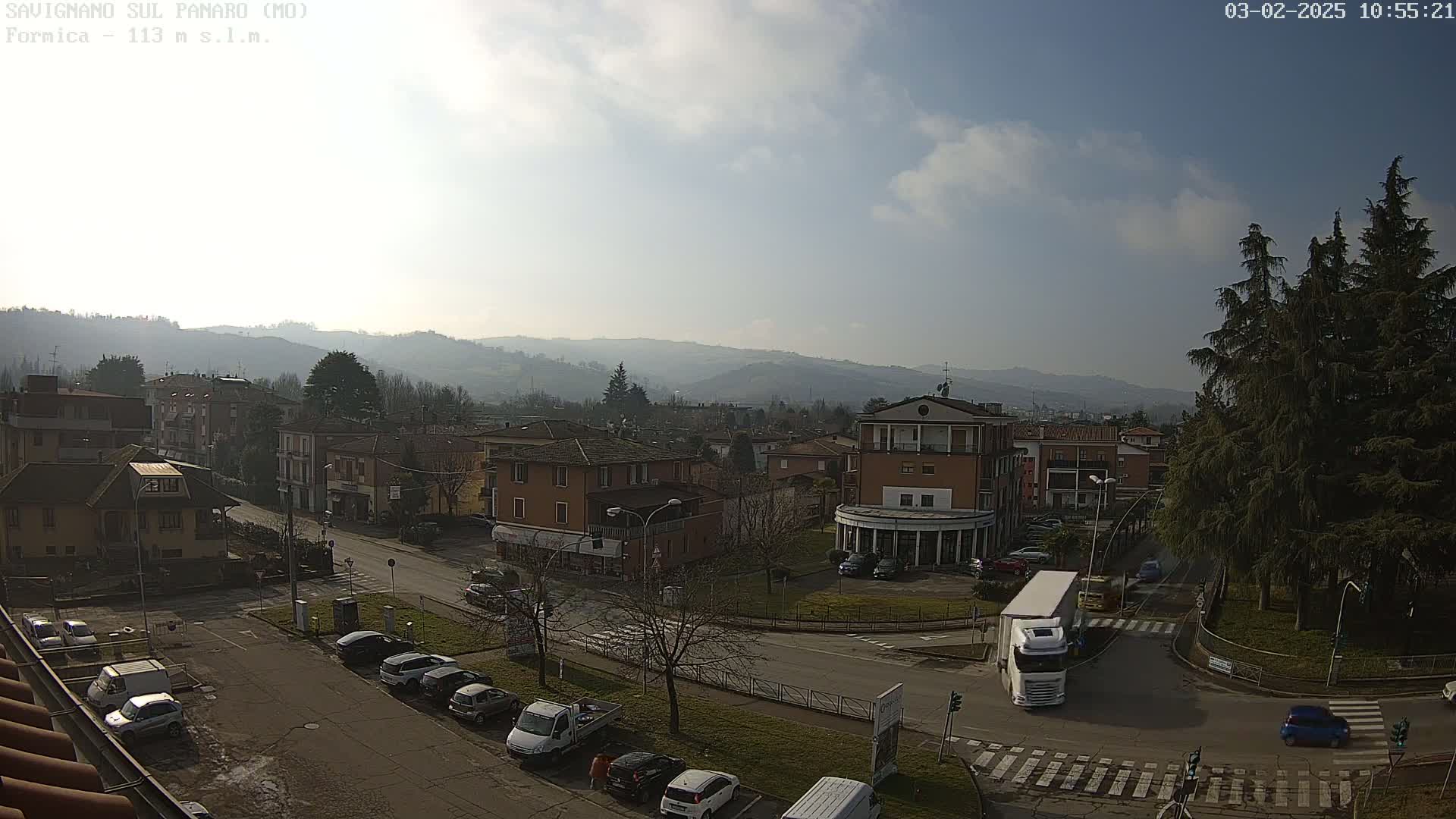 A sunny day reveals a town nestled in a valley, with cars parked along streets and a large truck at an intersection, surrounded by buildings and hills in the background.