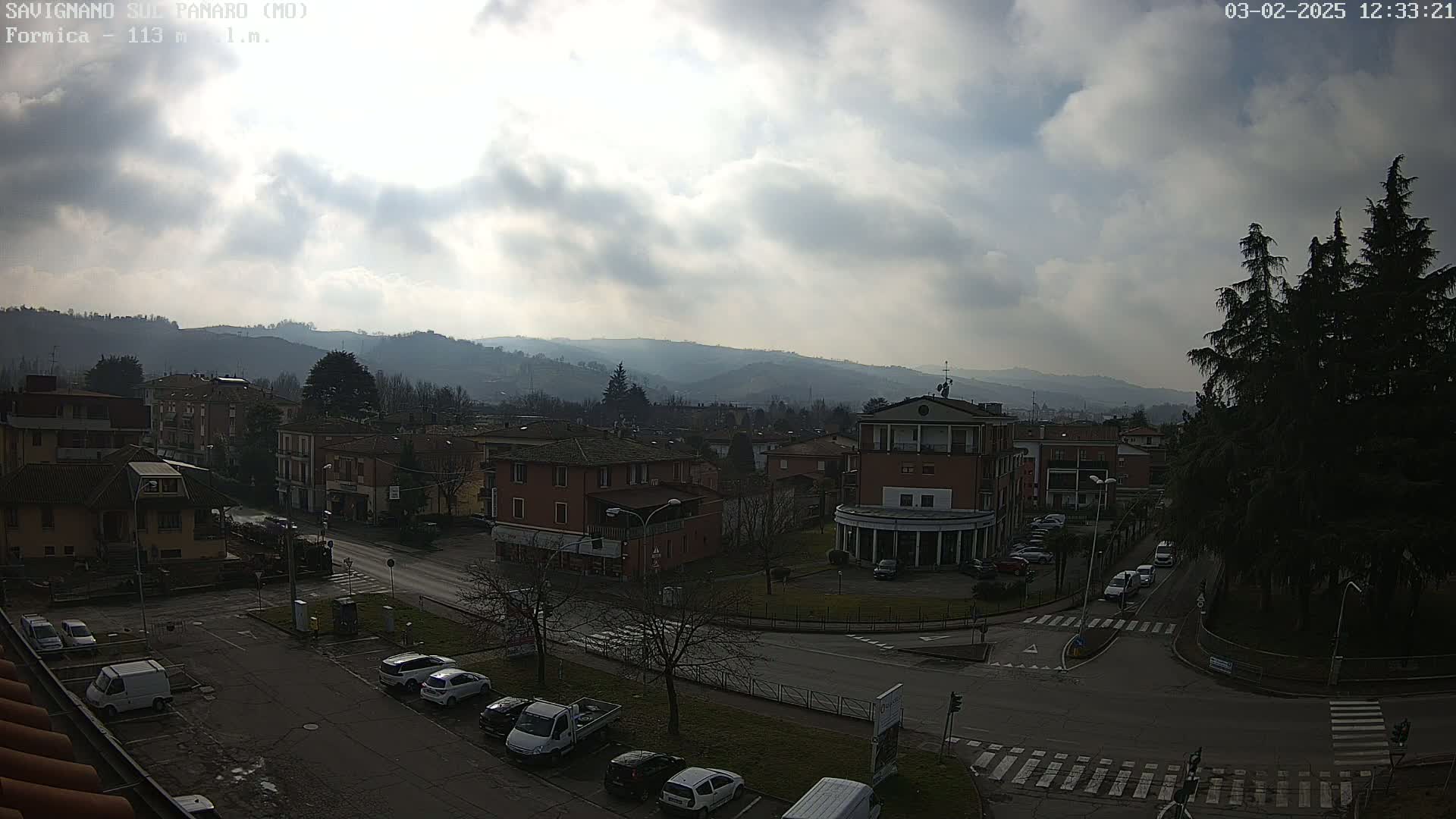 A partly cloudy day reveals a view of a town nestled among hills, with cars parked along streets and a roundabout in the foreground.