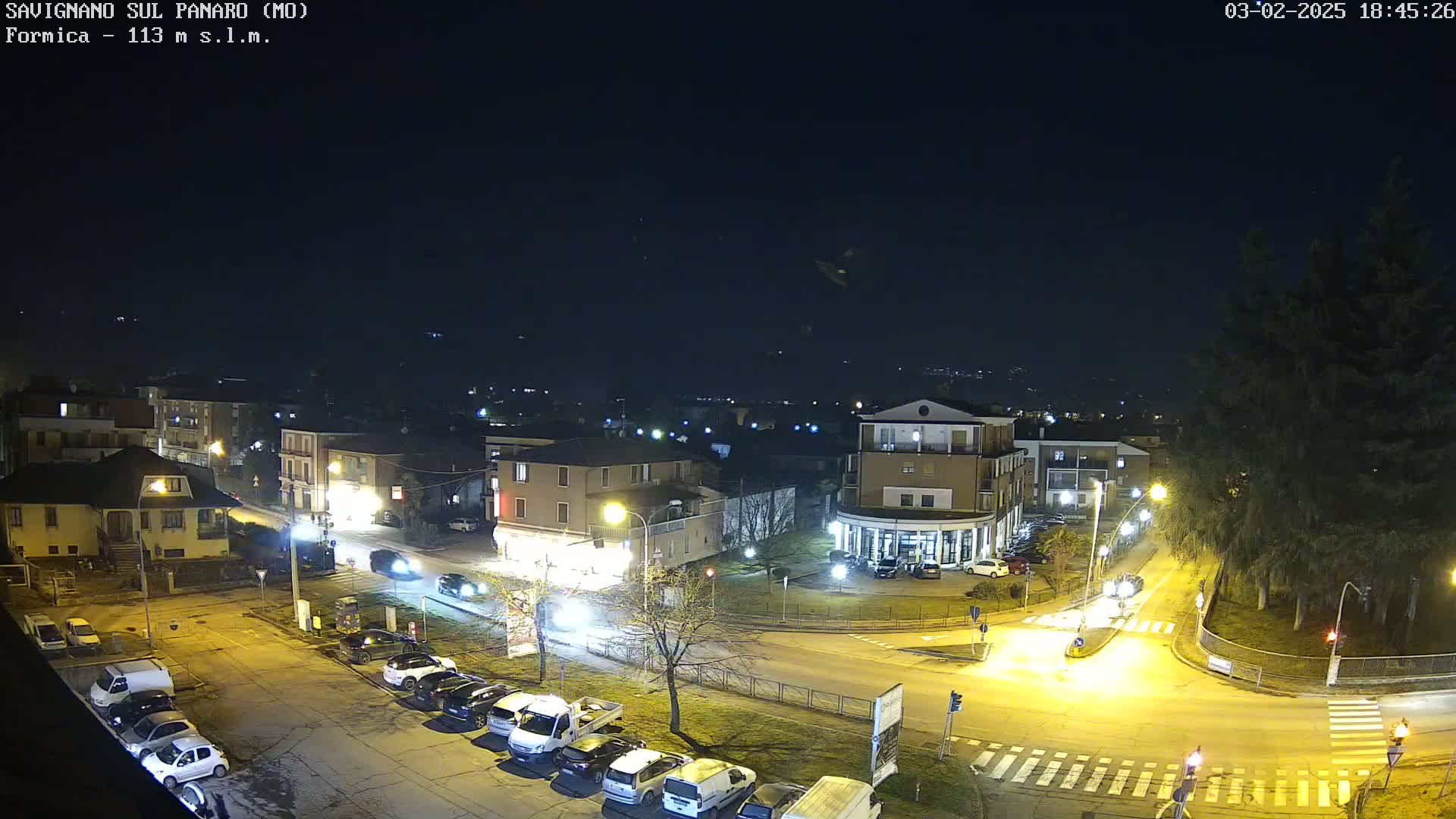 A nighttime aerial view of a town's street intersection with parked cars and buildings illuminated under a clear, dark sky.