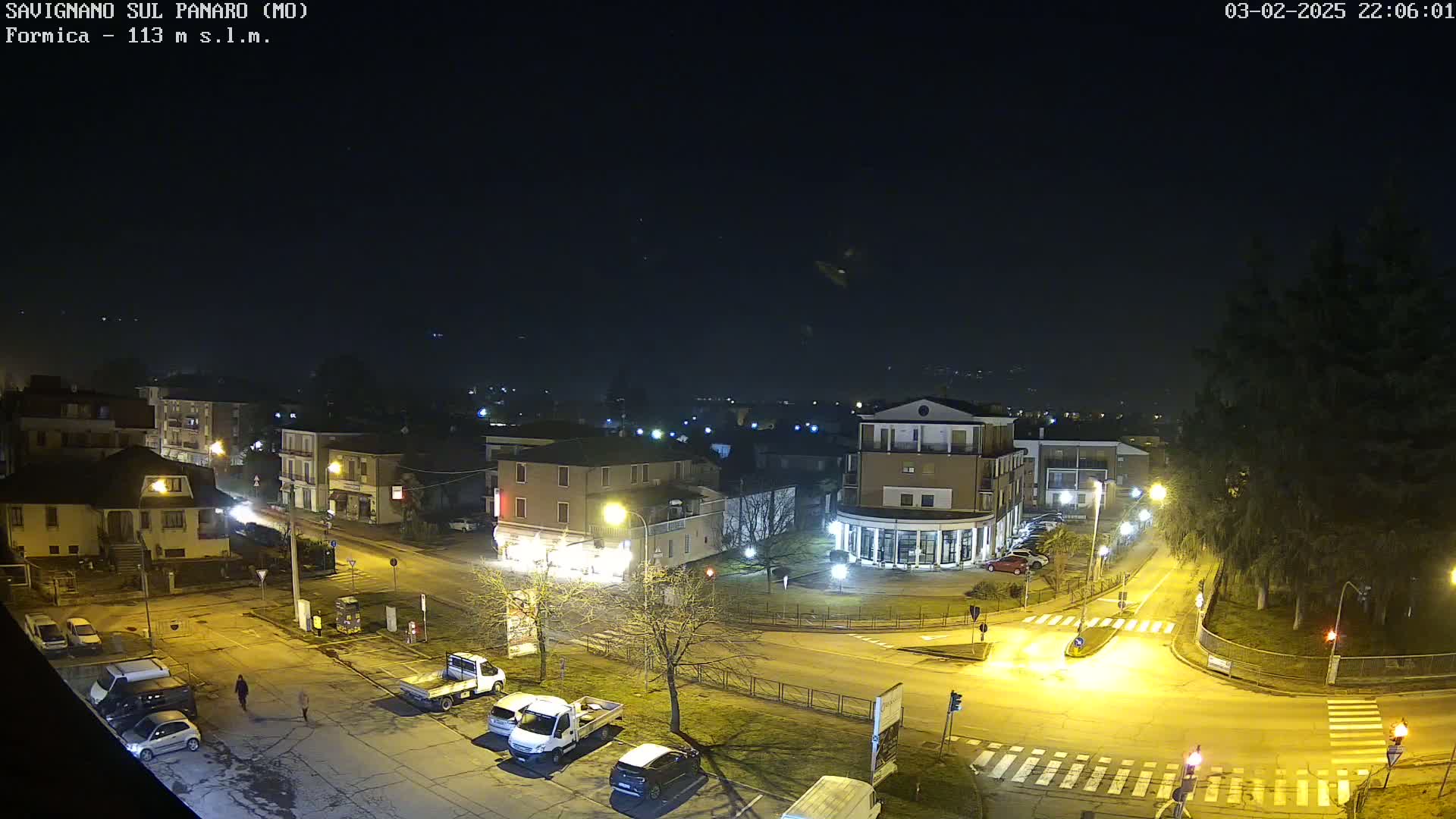 A nighttime aerial view of a town square with several buildings, parked cars, and a few pedestrians, under a clear, dark sky.