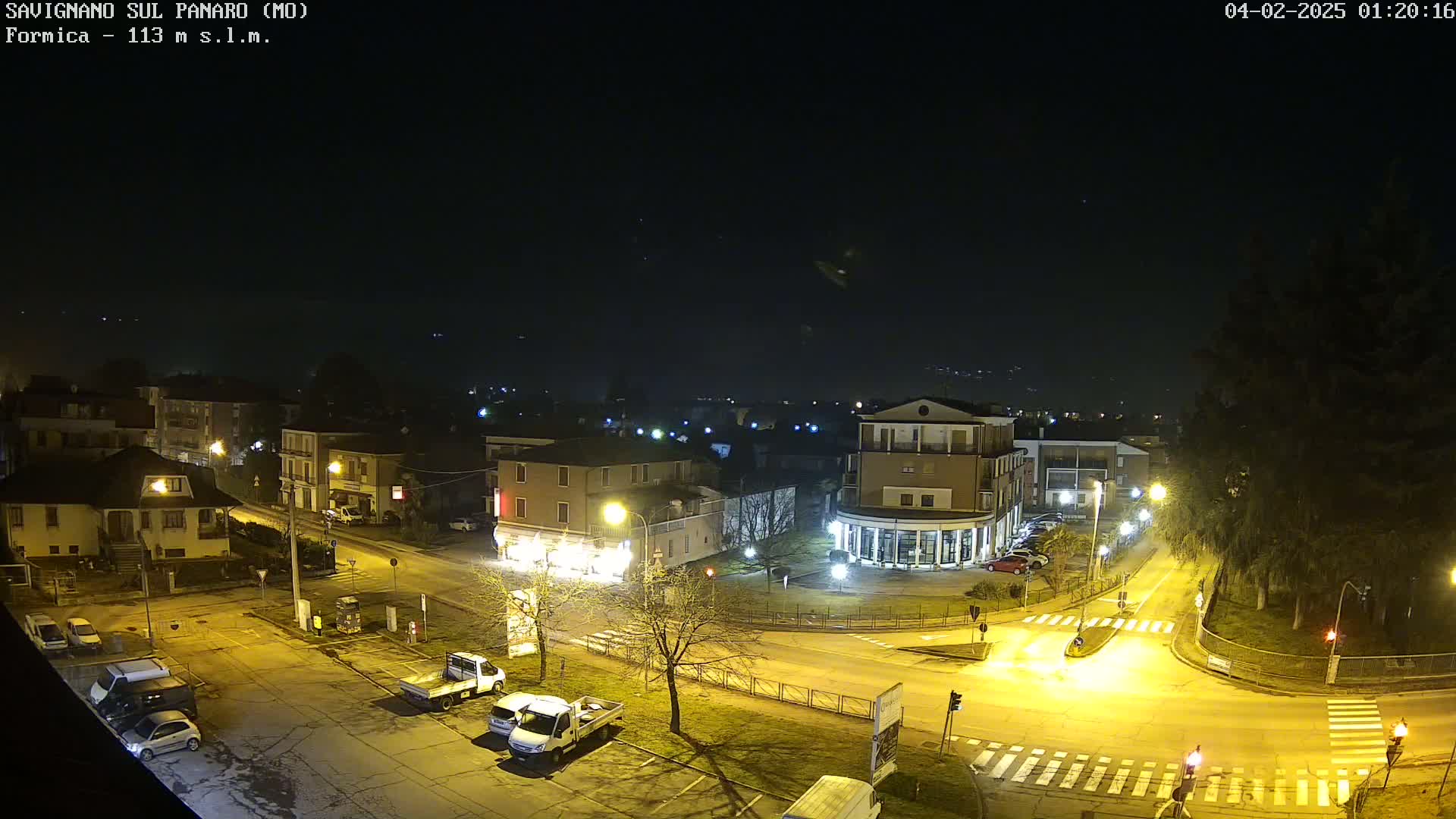A nighttime, overhead view of a town square with several buildings, parked cars, and a few vehicles on the roads, under a clear, dark sky.