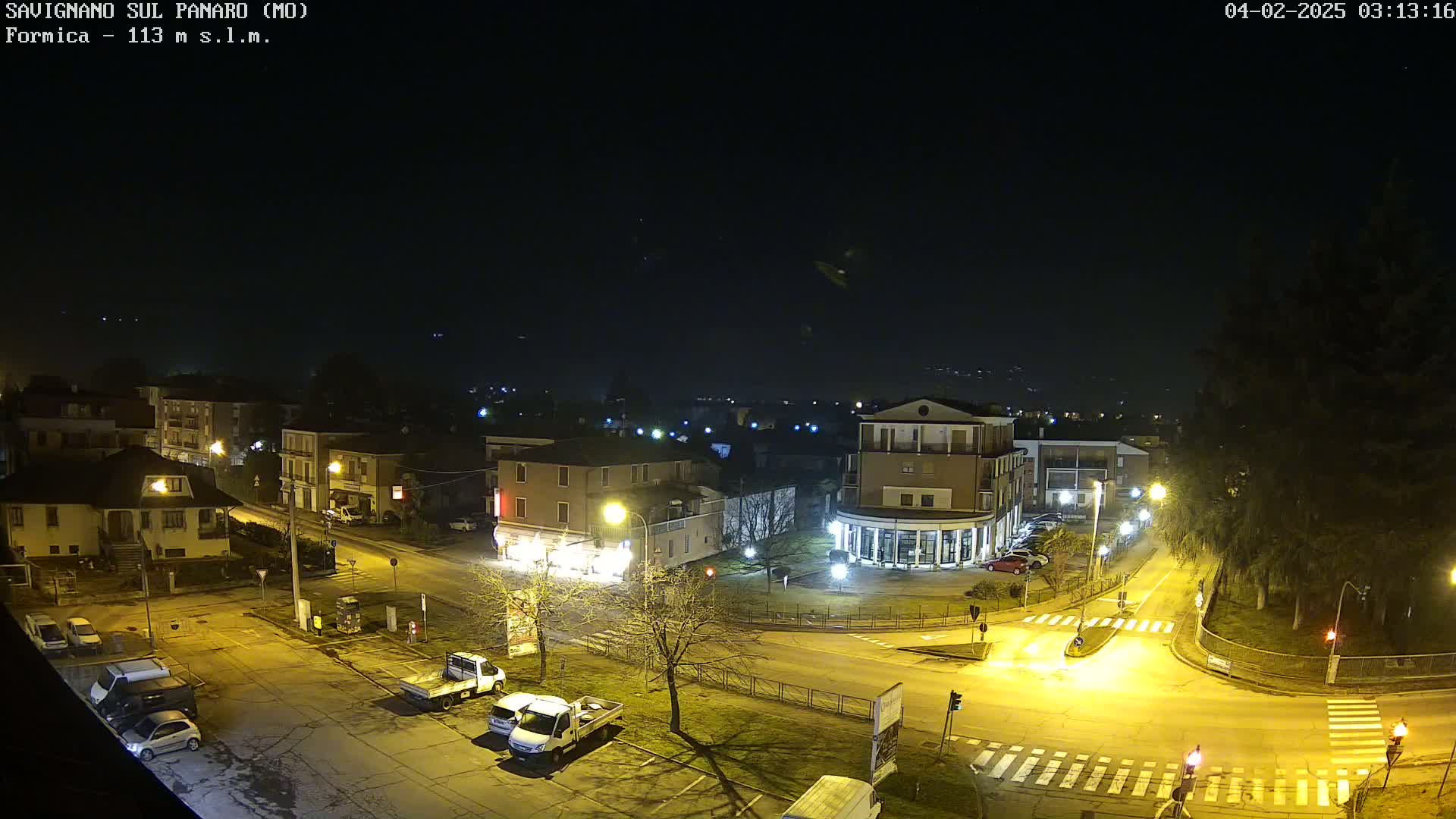 A nighttime view of a town square with parked cars and illuminated buildings under a clear, dark sky.