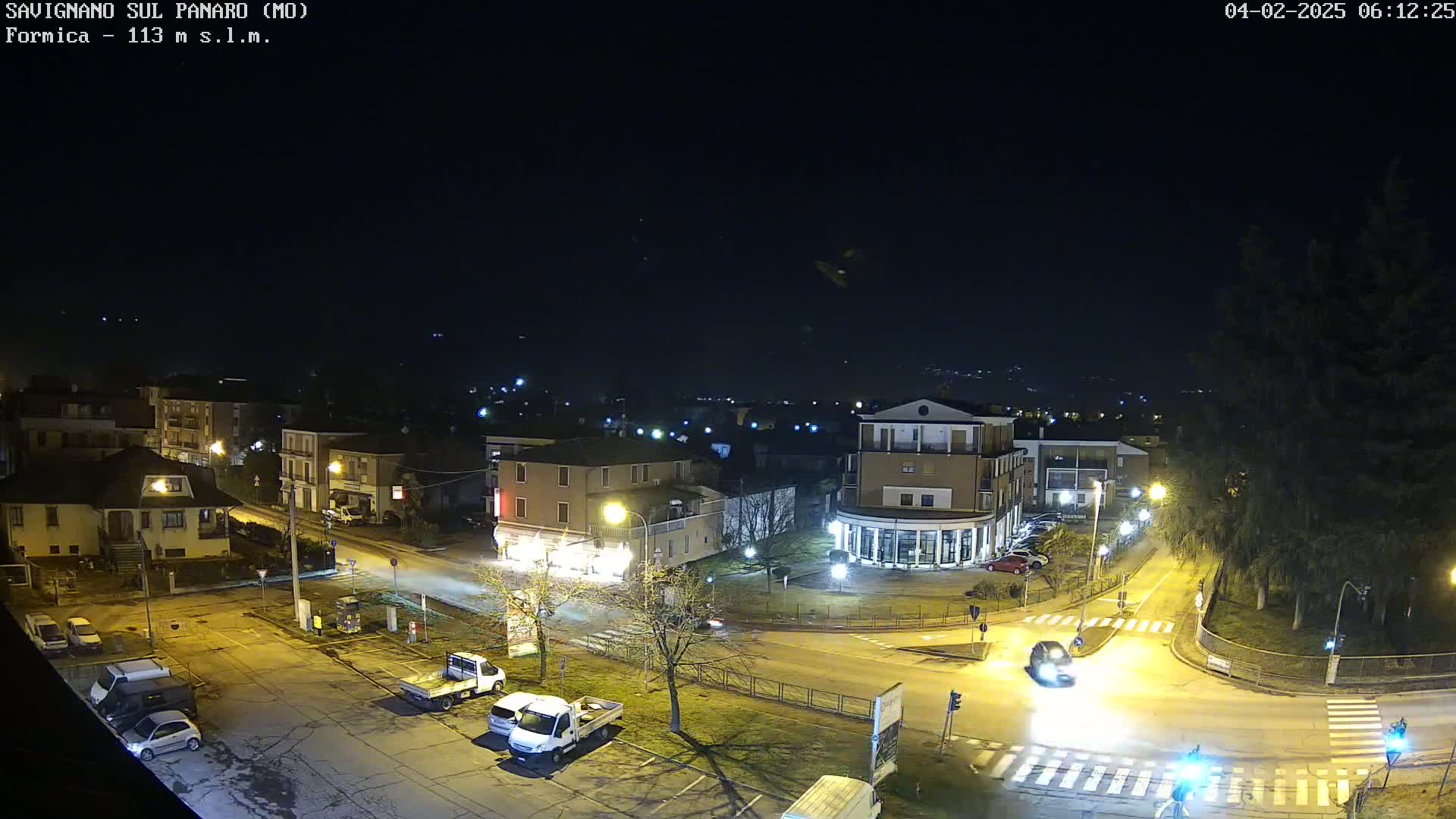 A nighttime aerial view of a town square with several cars and buildings illuminated by streetlights under a clear, dark sky.