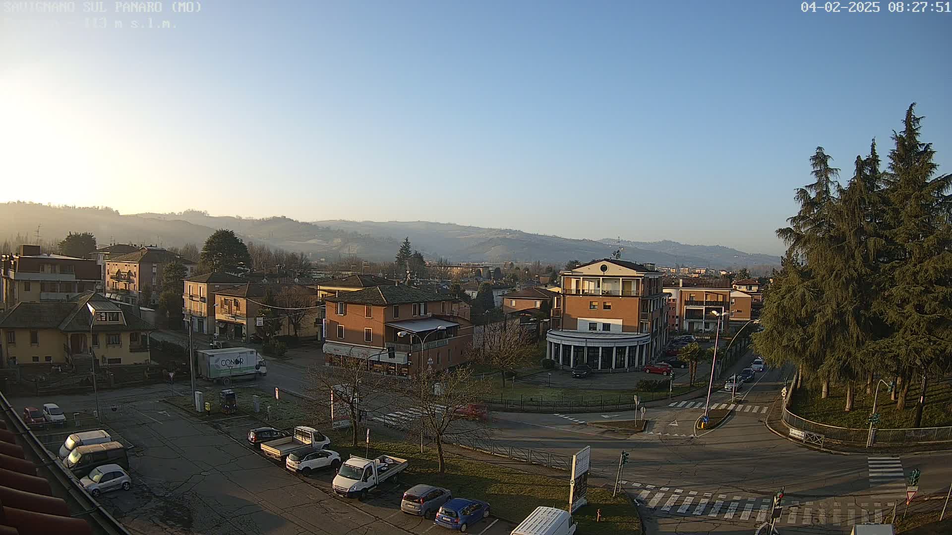 A clear, sunny day reveals a town nestled in a valley, with cars parked along the streets and a few vehicles on the road, surrounded by hills in the distance.