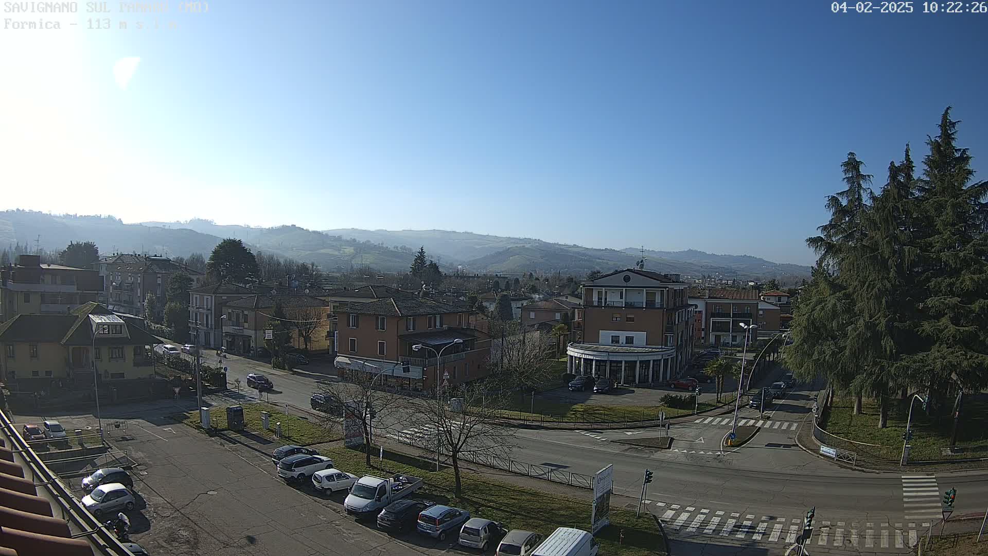A sunny day reveals a town nestled in a valley, with cars parked along streets and buildings of varying heights, and tall evergreens near a road intersection.