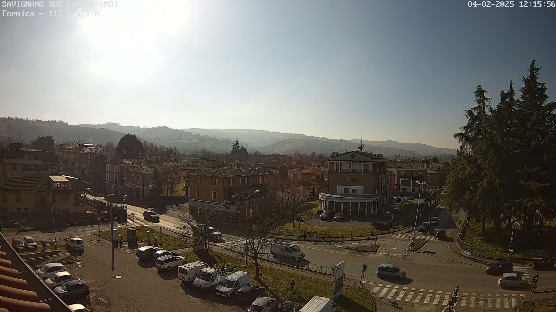 A sunny day reveals a town nestled in a valley, with cars parked along streets and a few vehicles driving through a small roundabout.