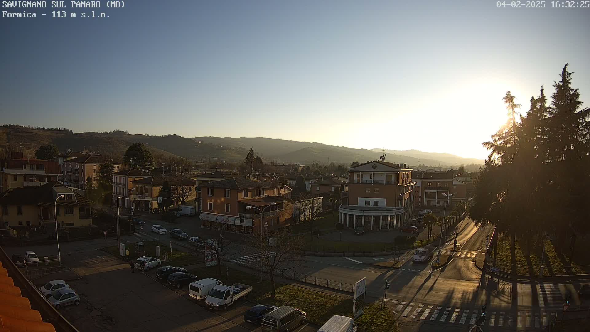 An elevated view of a town at sunset, showing buildings, streets, parked cars, and trees, under a clear sky.