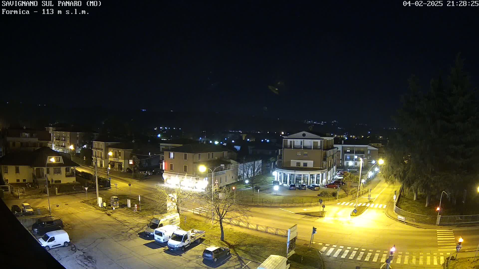 A nighttime aerial view of a town square with several buildings, vehicles, and streetlights, under a clear, dark sky.