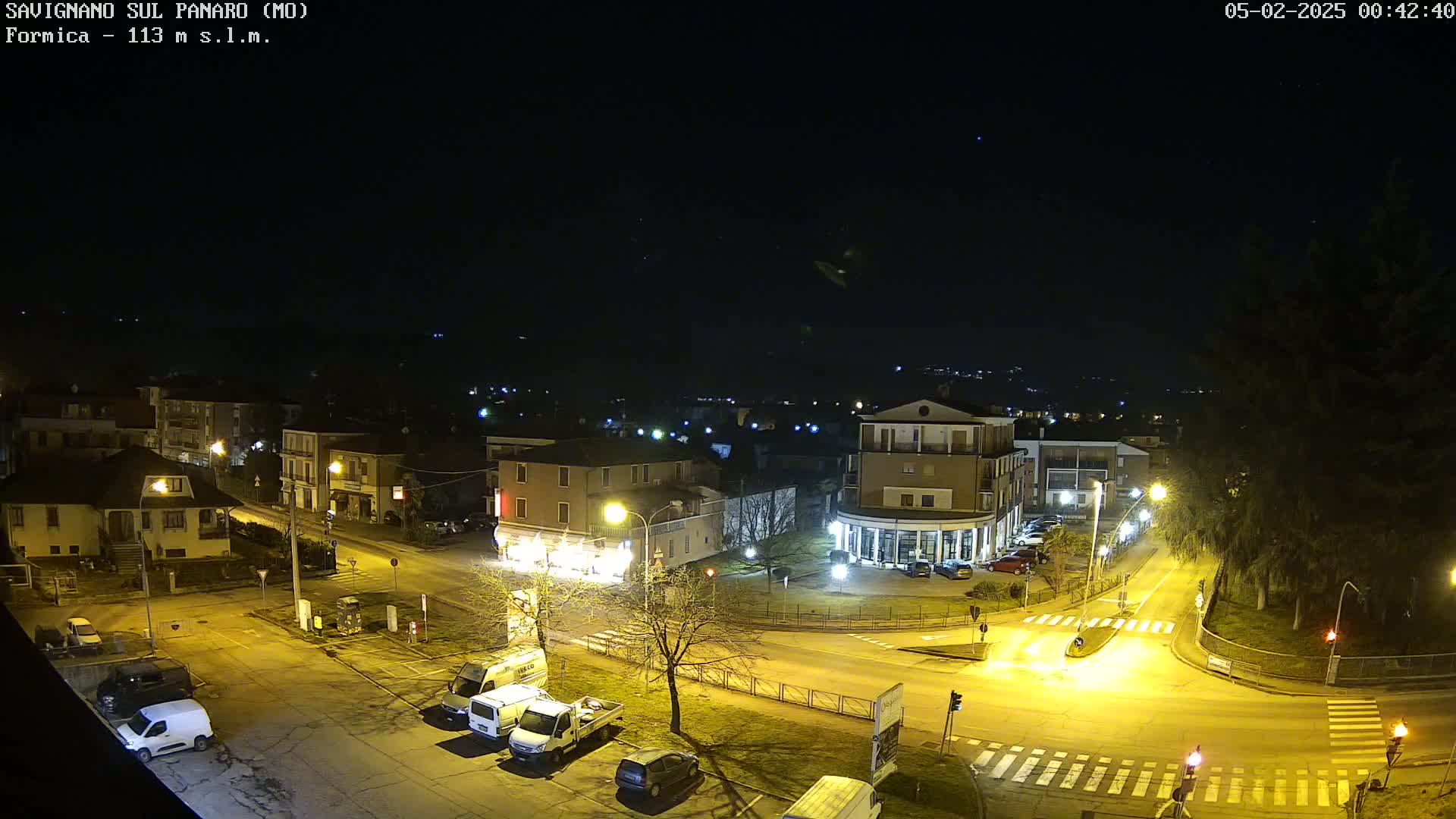 A nighttime aerial view of a town square with several buildings and cars parked along the streets under a clear, dark sky.