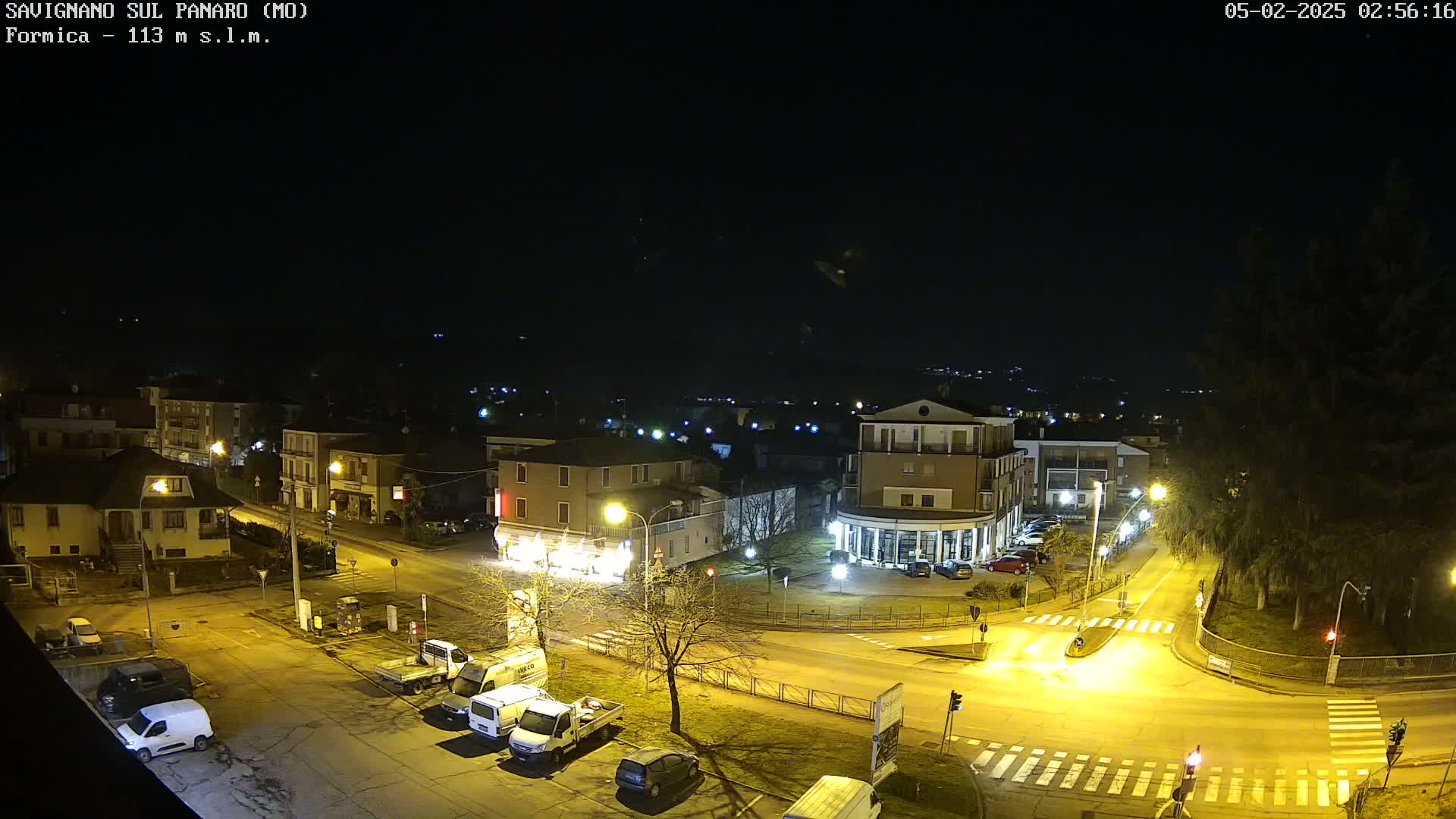 A nighttime, overhead view of a town square with buildings, parked cars, and streetlights illuminating the roads and surrounding area under a clear, dark sky.