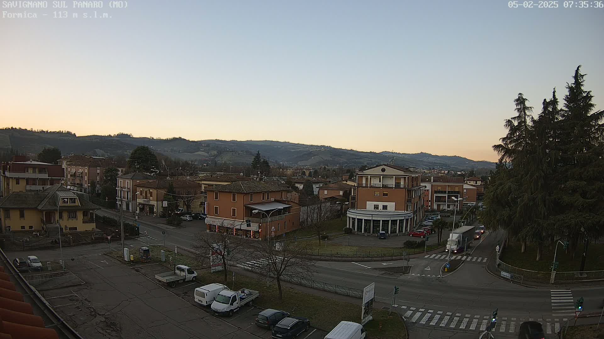 A high-angle, long shot shows a town nestled in a valley at sunrise under a clear sky, with cars parked along roads and a large truck at an intersection.