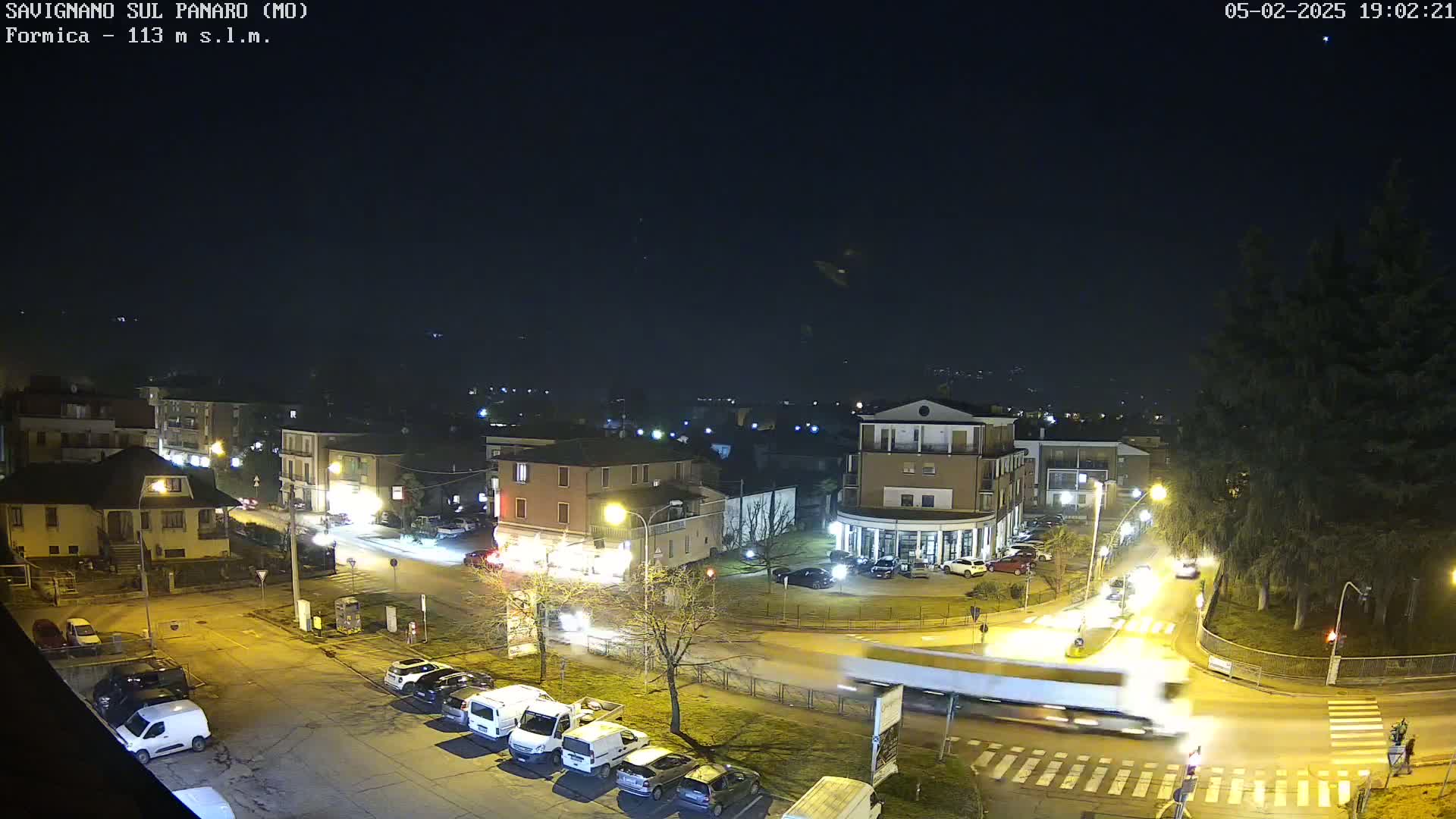 A nighttime, high-angle view of a town square with cars parked and driving, streetlights illuminating the area, and a large truck moving through the intersection.