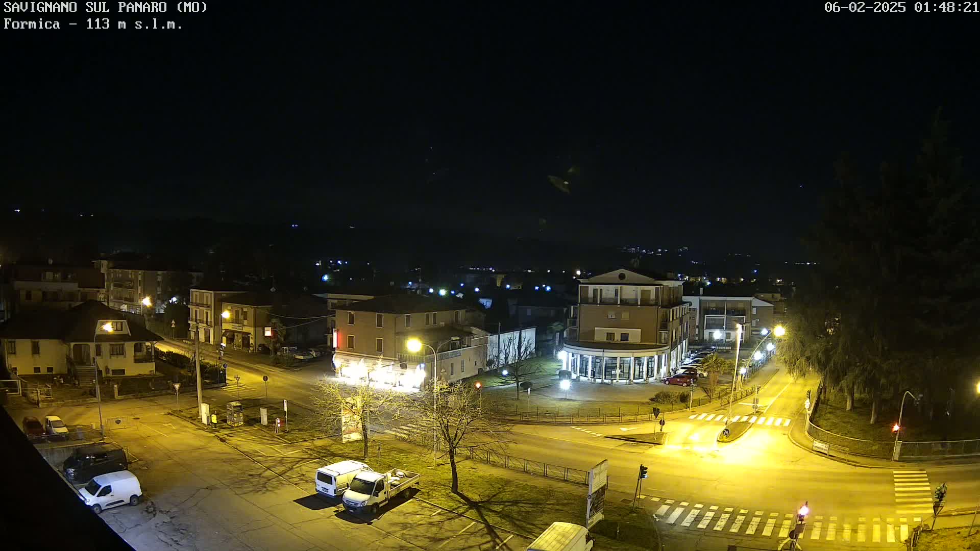 A nighttime aerial view of a town square with buildings, streets, and parked vehicles under a clear, dark sky.