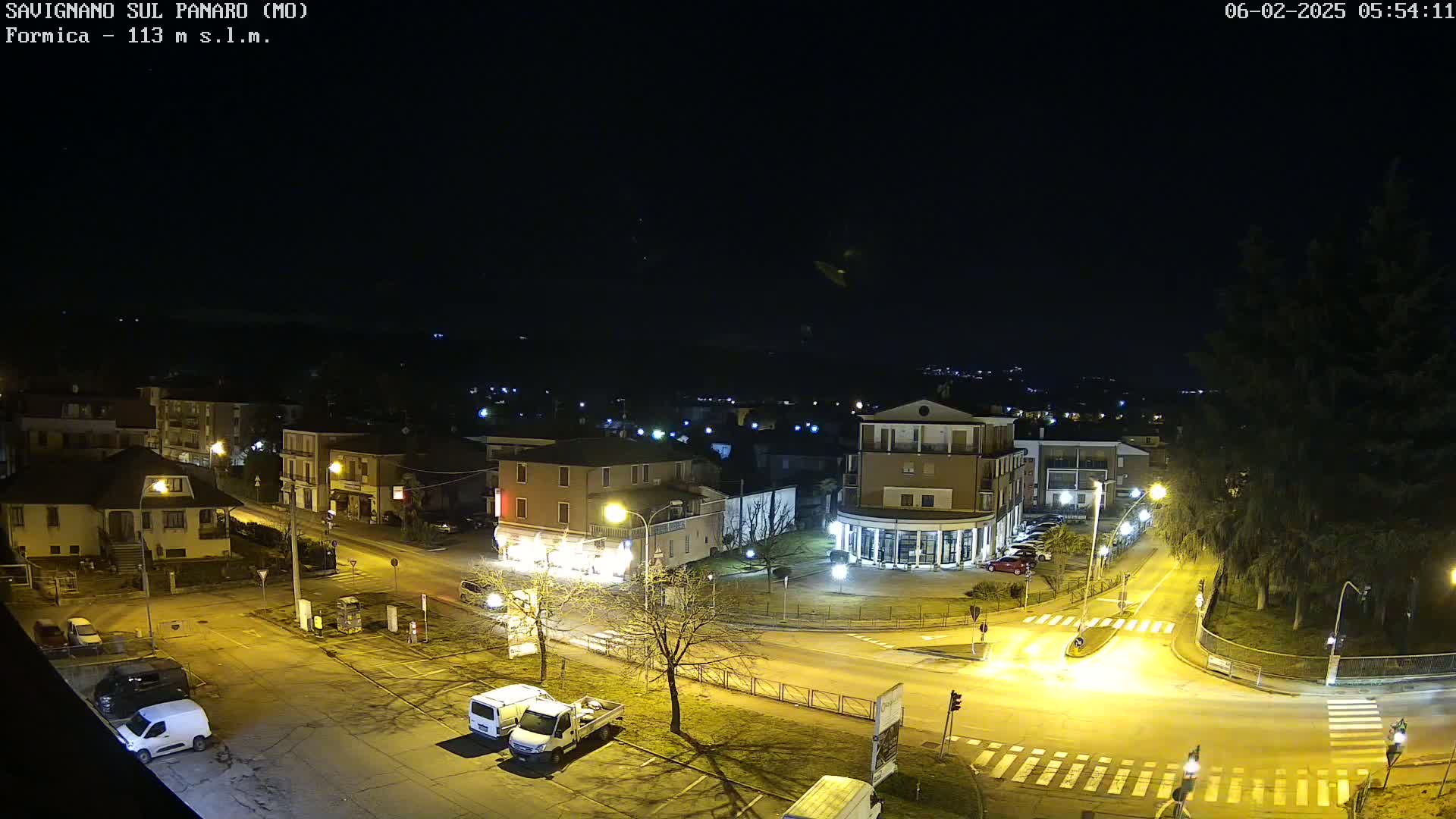 A nighttime, overhead view of a town square with buildings, streets, and parked cars, illuminated by streetlights.