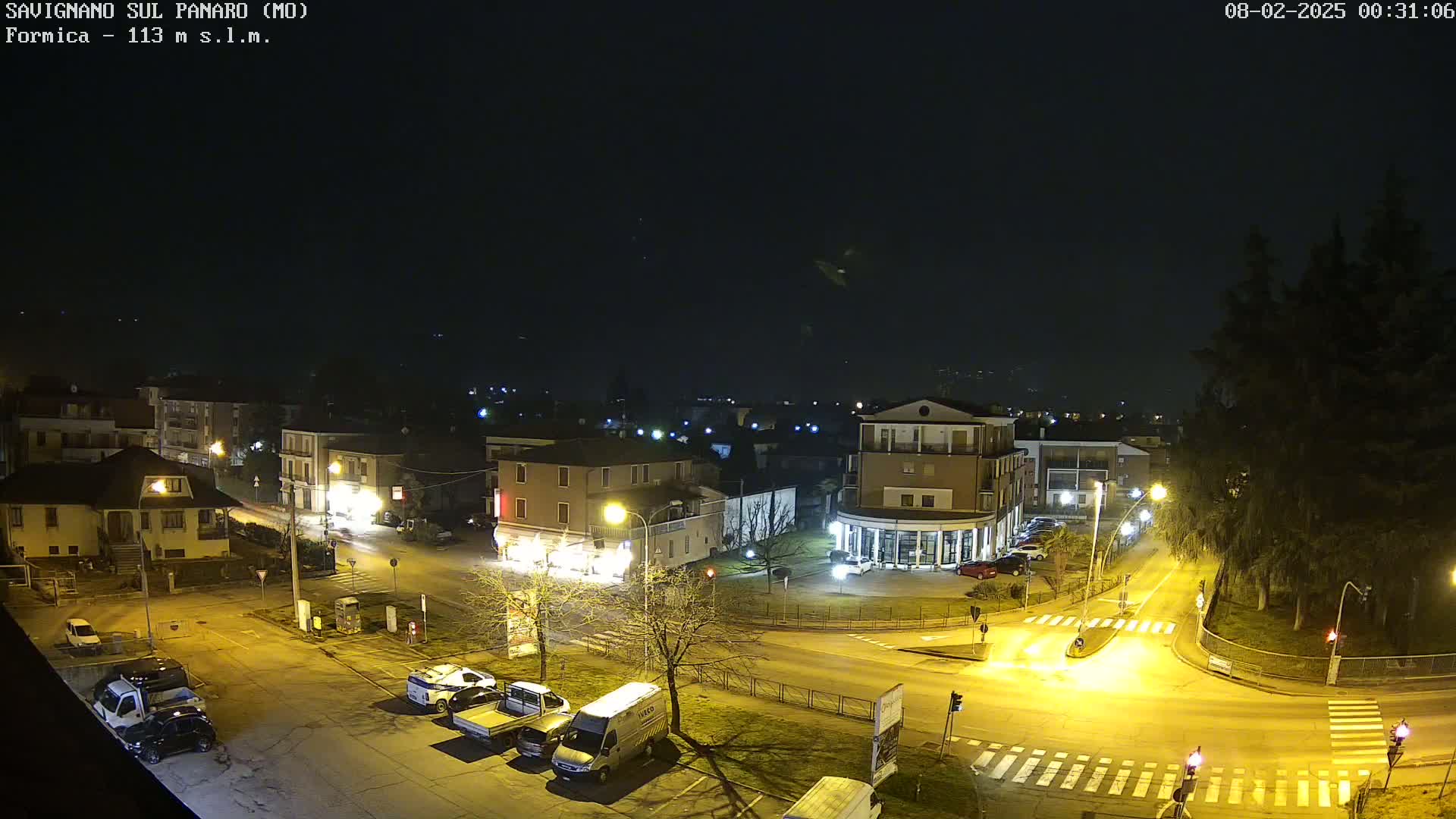 A nighttime, overhead view of a town square with several buildings, parked vehicles, and streetlights illuminating the scene under a dark sky.