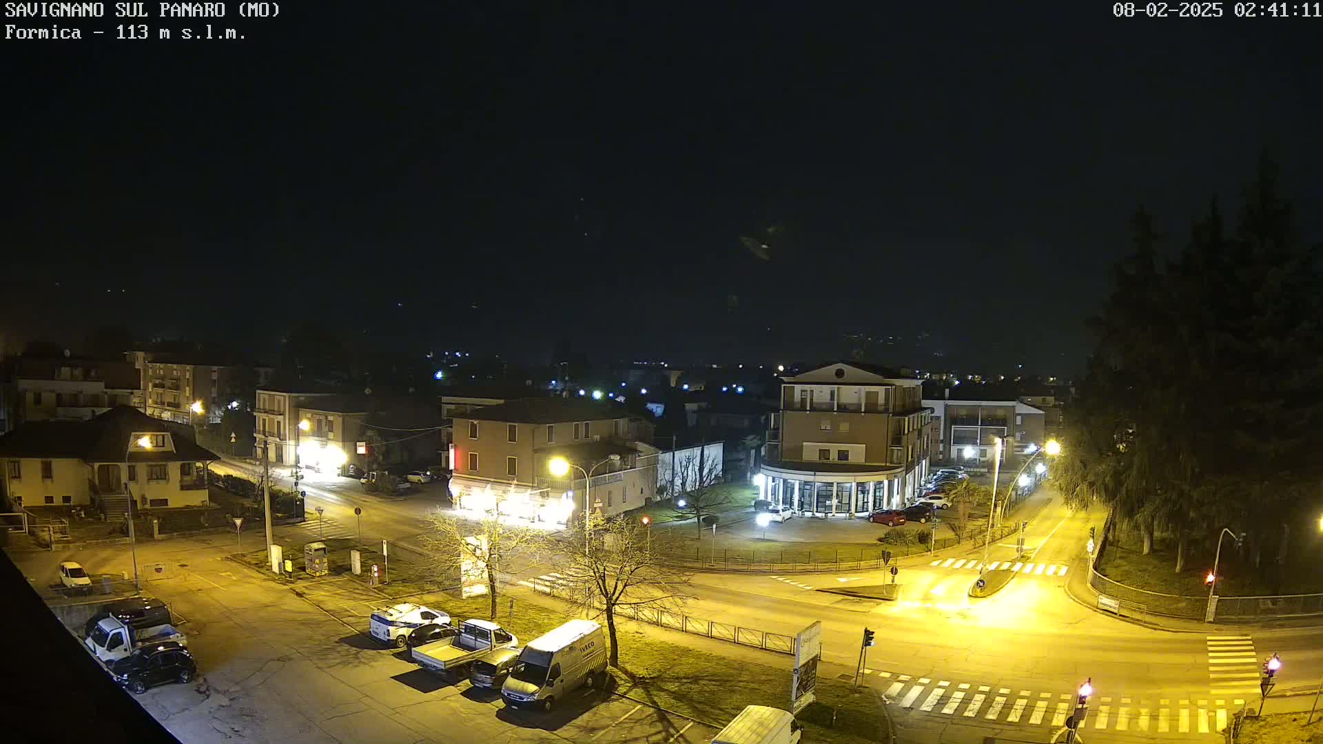 A nighttime aerial view of a town square with parked cars and streetlights illuminating the streets and buildings under a clear, dark sky.