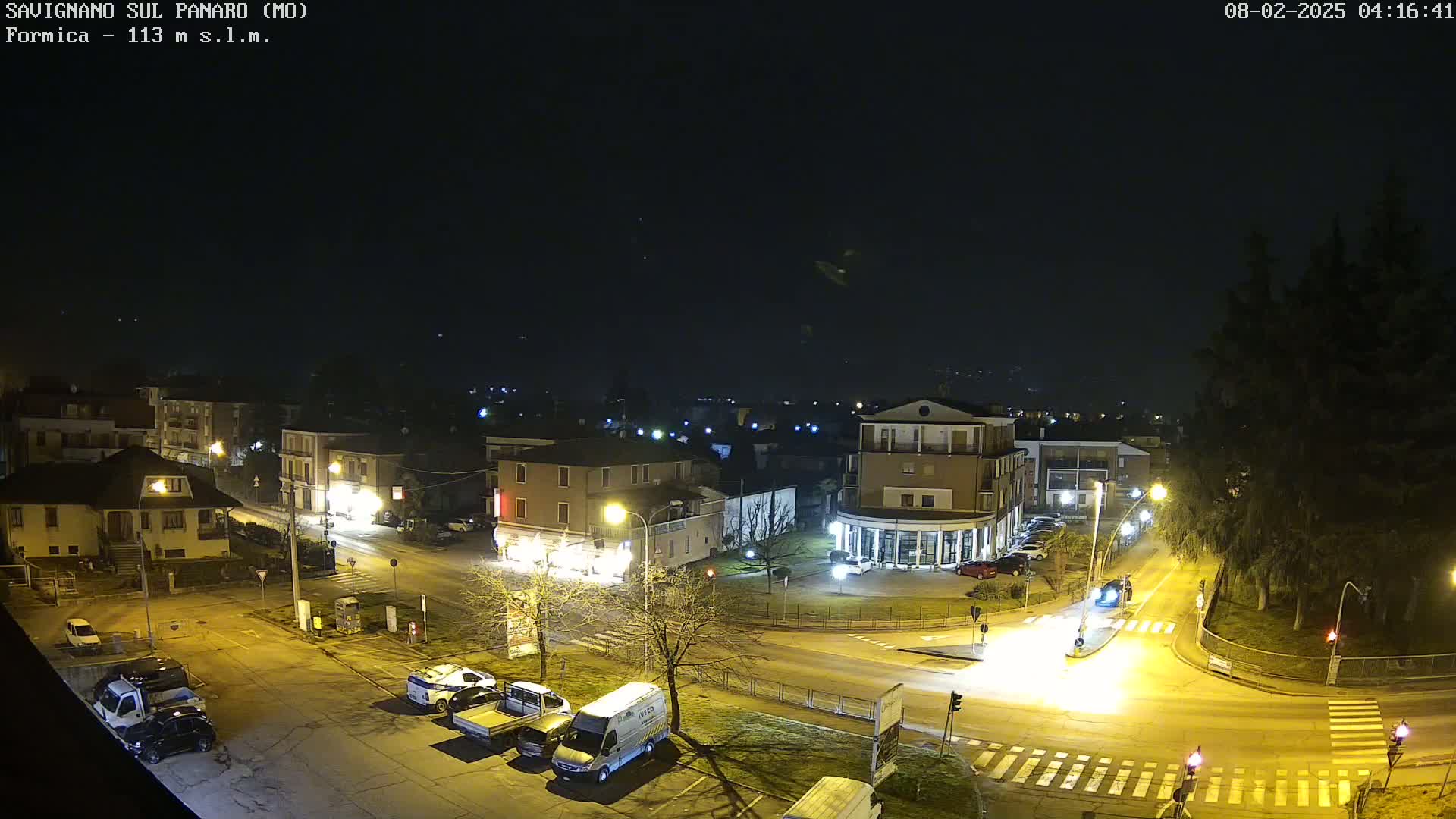 A nighttime aerial view of a town square with several buildings, vehicles, and streetlights under a clear, dark sky.