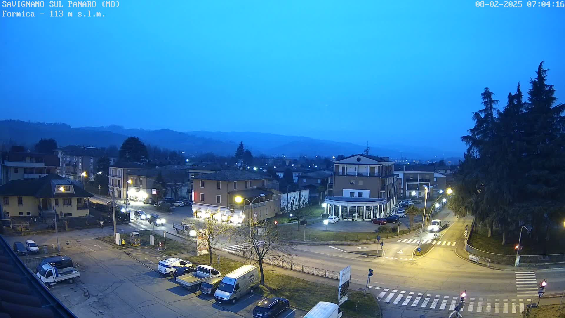 A twilight view of a town square with parked cars and buildings, under a clear blue-ish sky.