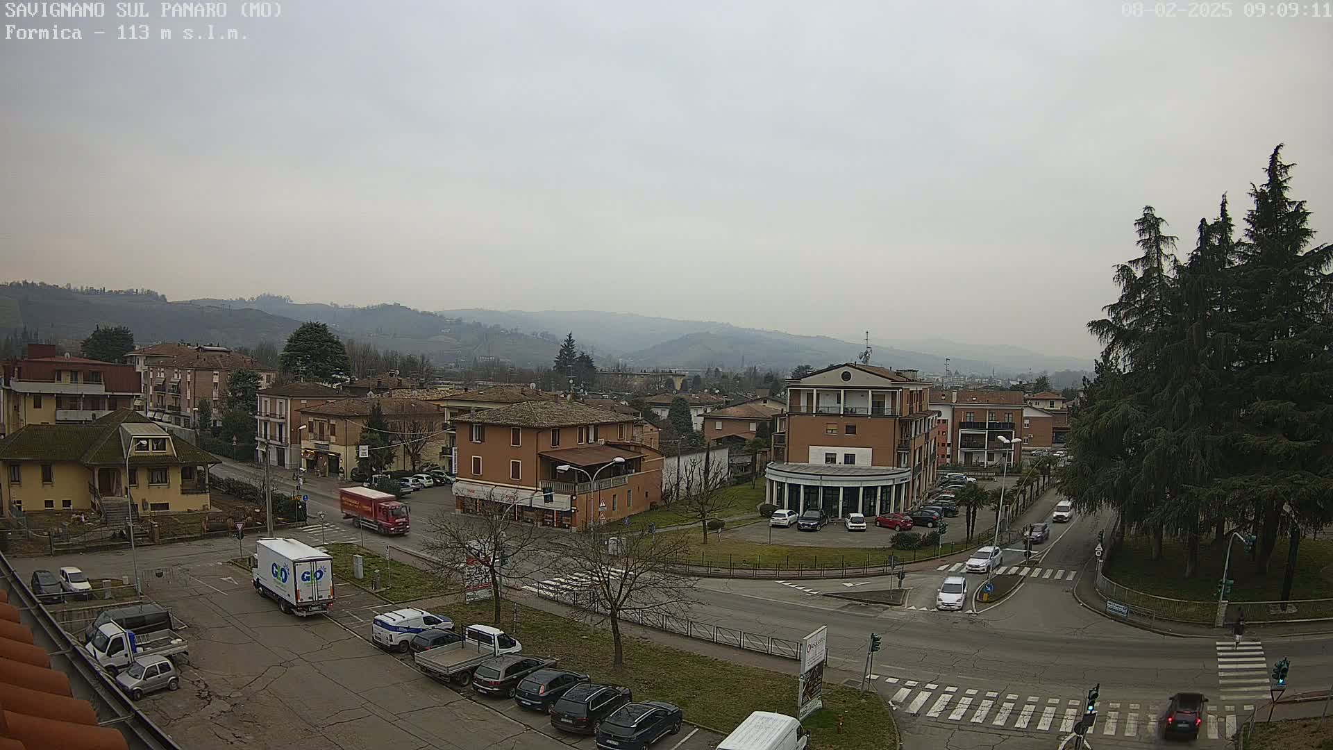 A mostly cloudy day reveals a small Italian town nestled in a valley, with several cars parked in lots and driving on streets surrounding a small roundabout.