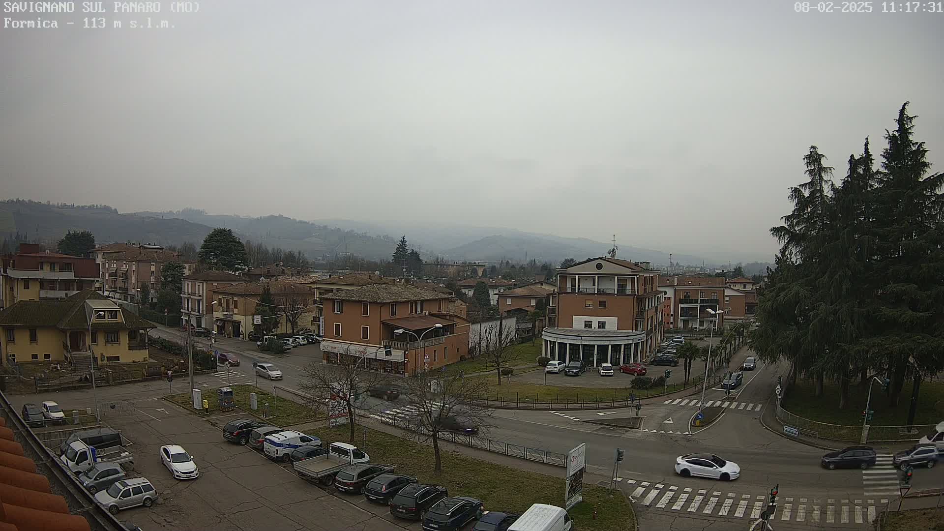 A hazy, overcast day reveals a town square in a hilly area, with several cars parked and driving near buildings and trees.