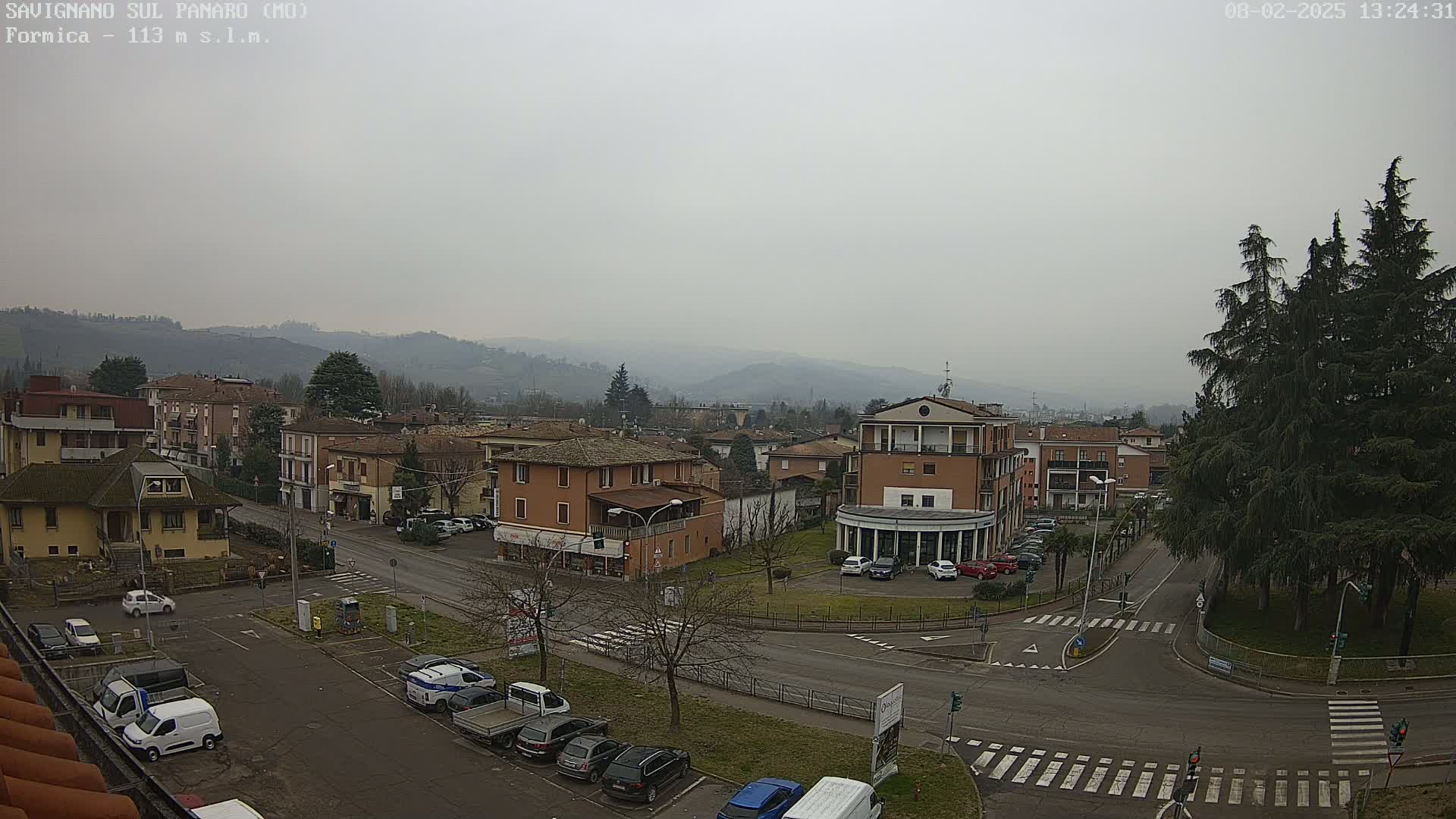 A hazy, overcast view of a town square with parked cars and buildings, surrounded by hills in the distance.