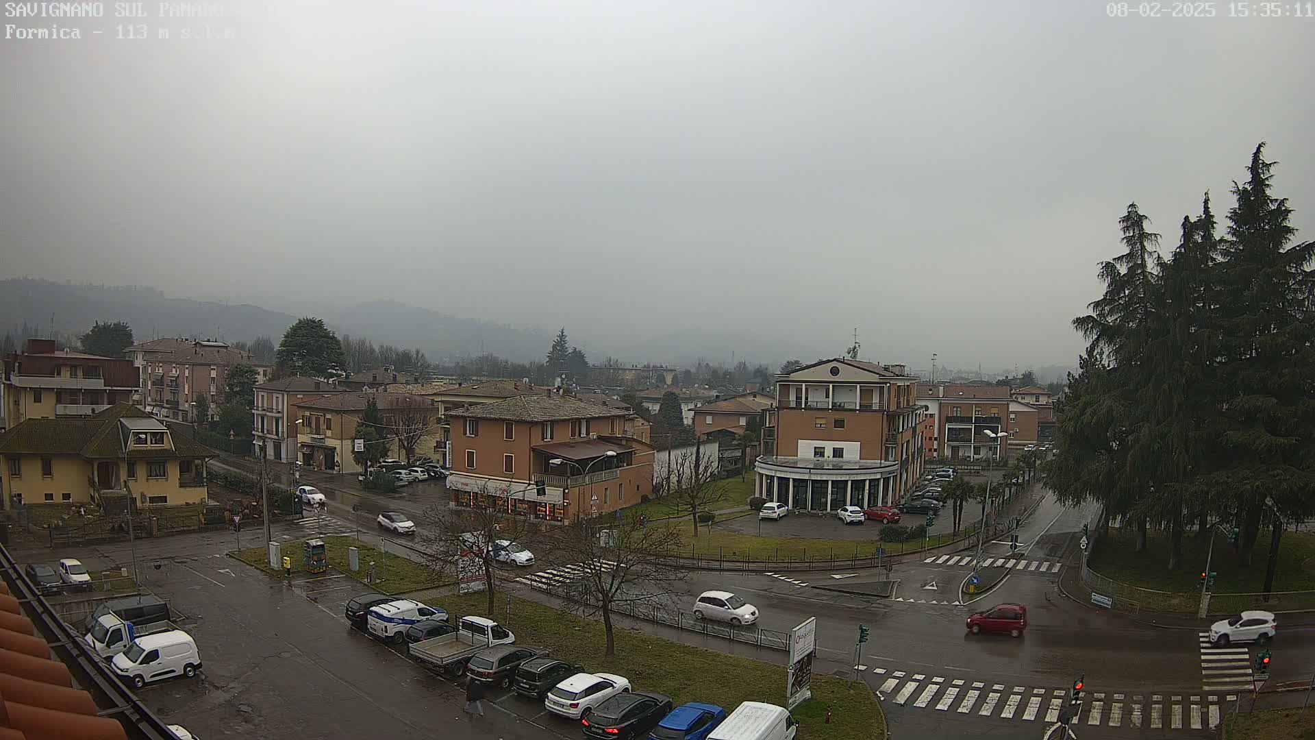 A view of a town square on an overcast day, showing several buildings, cars, and wet roads, with hills visible in the background.