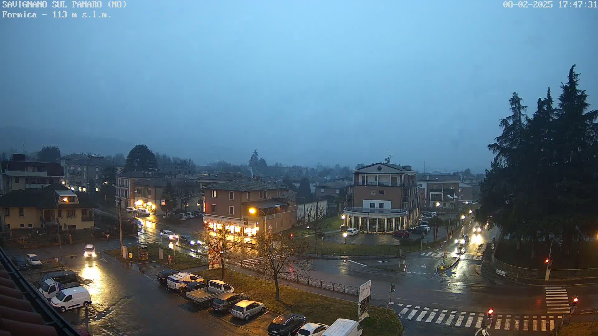 A rain-soaked town square at dusk, with cars parked and driving on wet streets under a hazy, overcast sky.