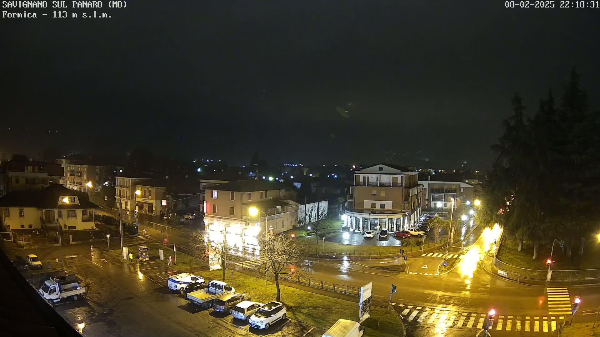 A rain-slicked nighttime street scene shows a town square with parked cars and buildings under a dark, cloudy sky.