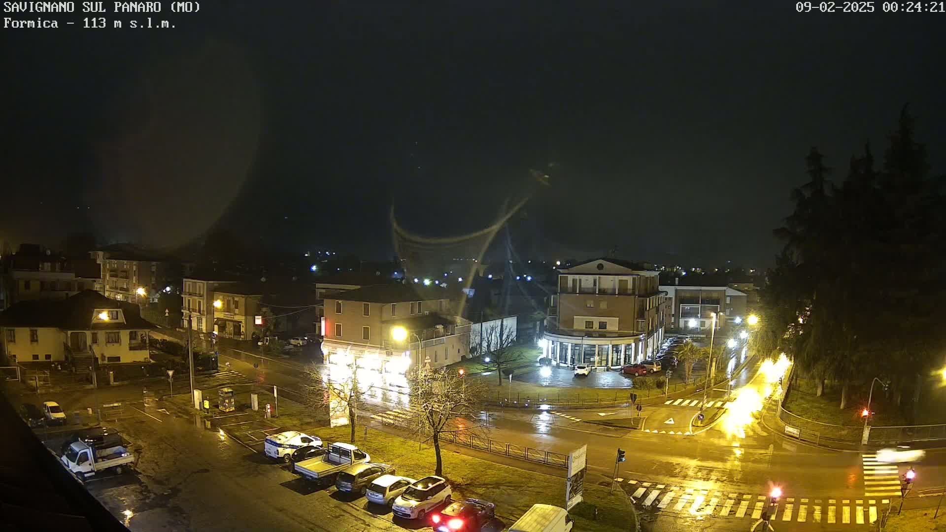 A nighttime view of a town square with wet streets and parked cars under a dark, possibly rainy sky.
