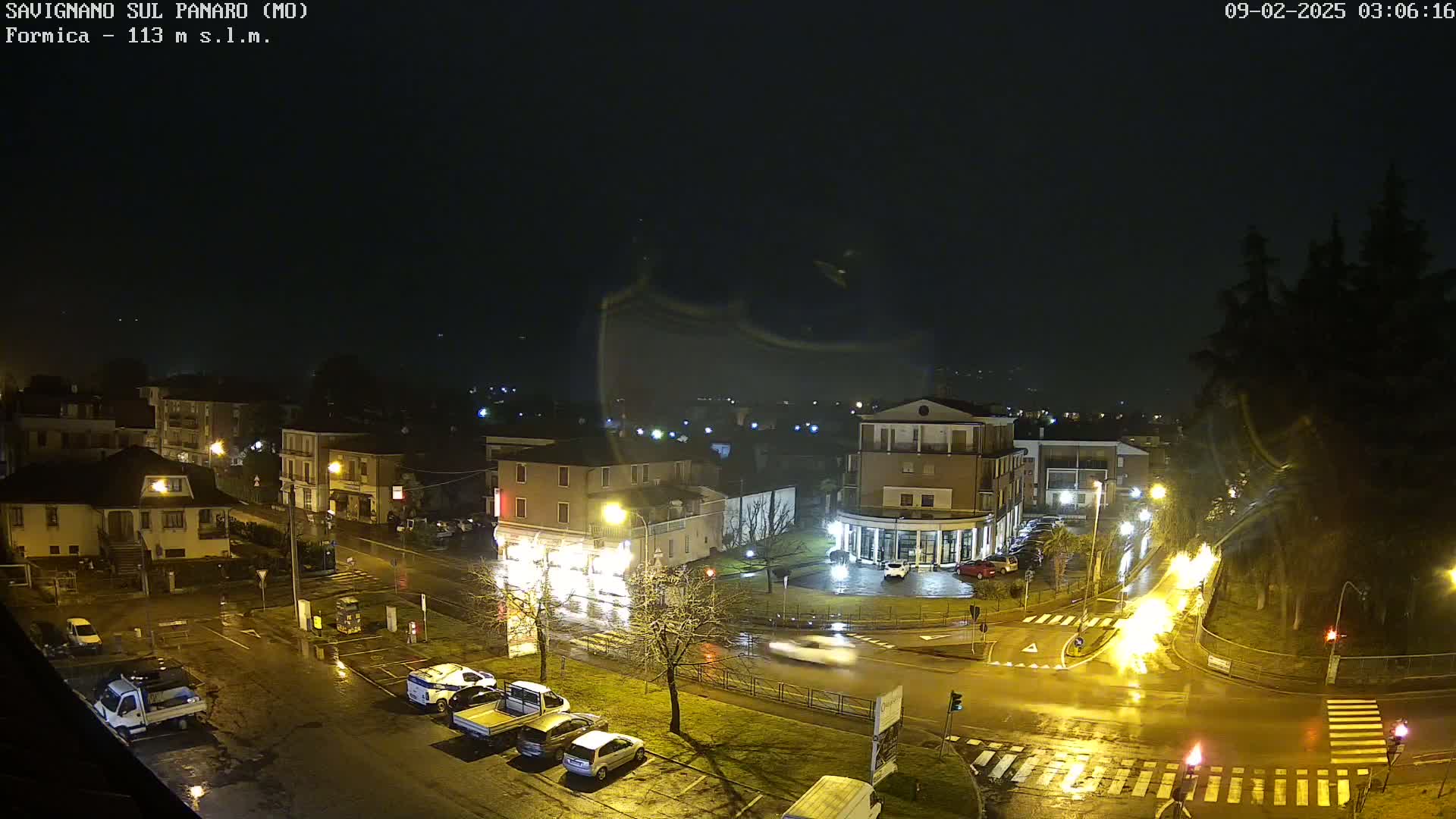 A nighttime, rain-slicked town square with several cars and buildings is seen from an elevated vantage point.