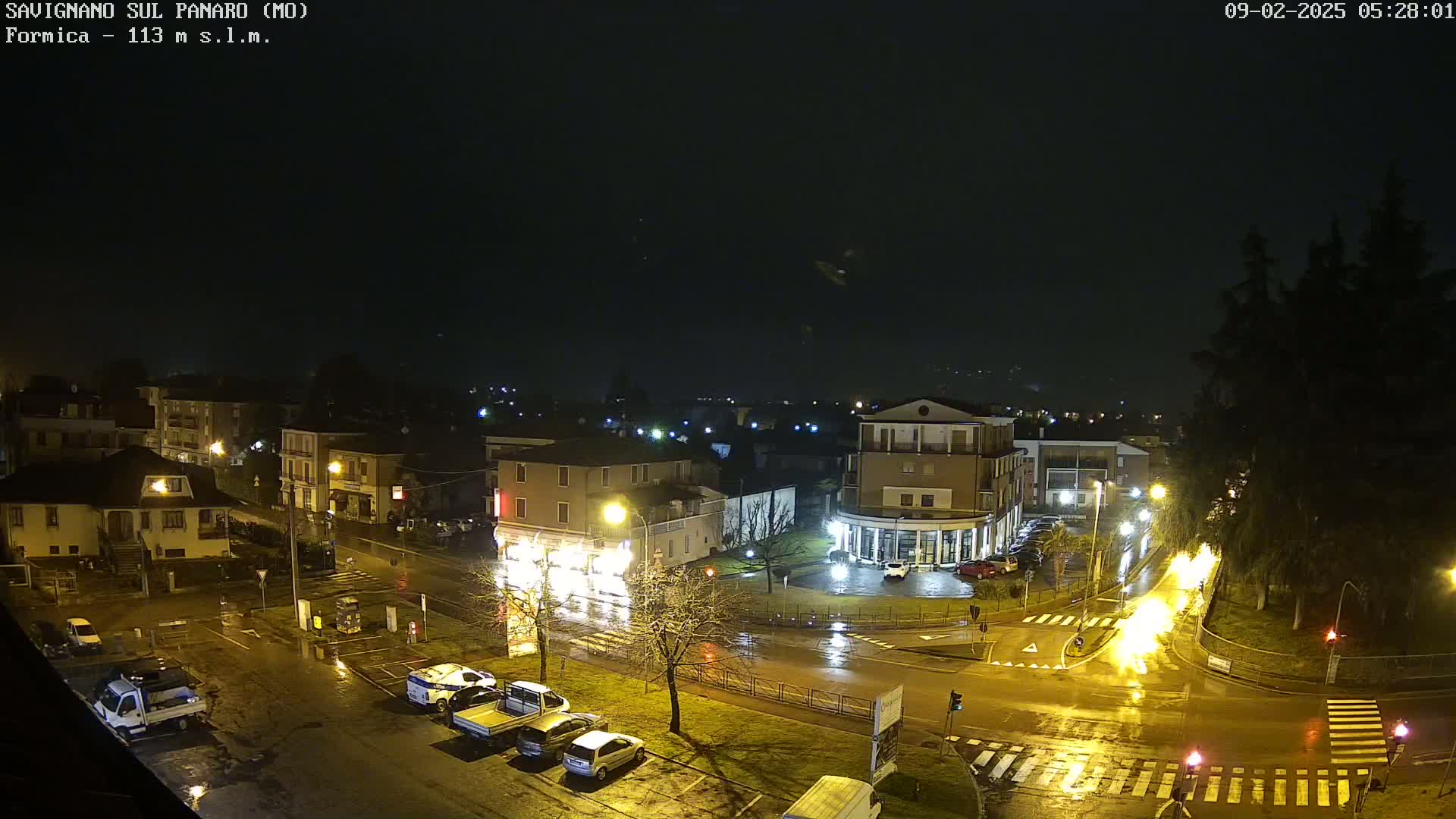 A wet, nighttime aerial view of a town square with buildings, parked cars, and streetlights illuminated during a light rain.