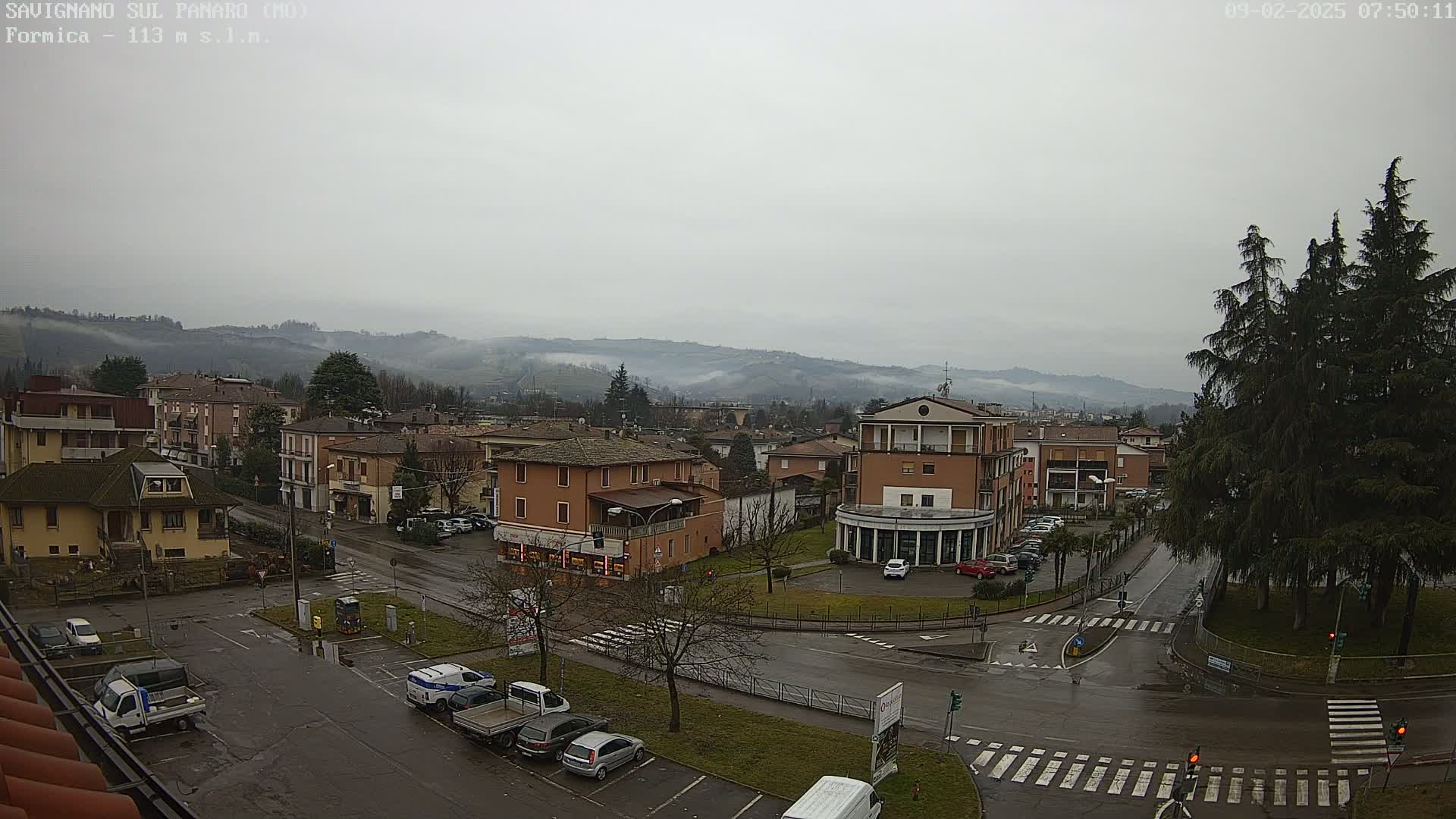 A wet, overcast town square in a valley, surrounded by low, fog-covered hills and tall evergreen trees.