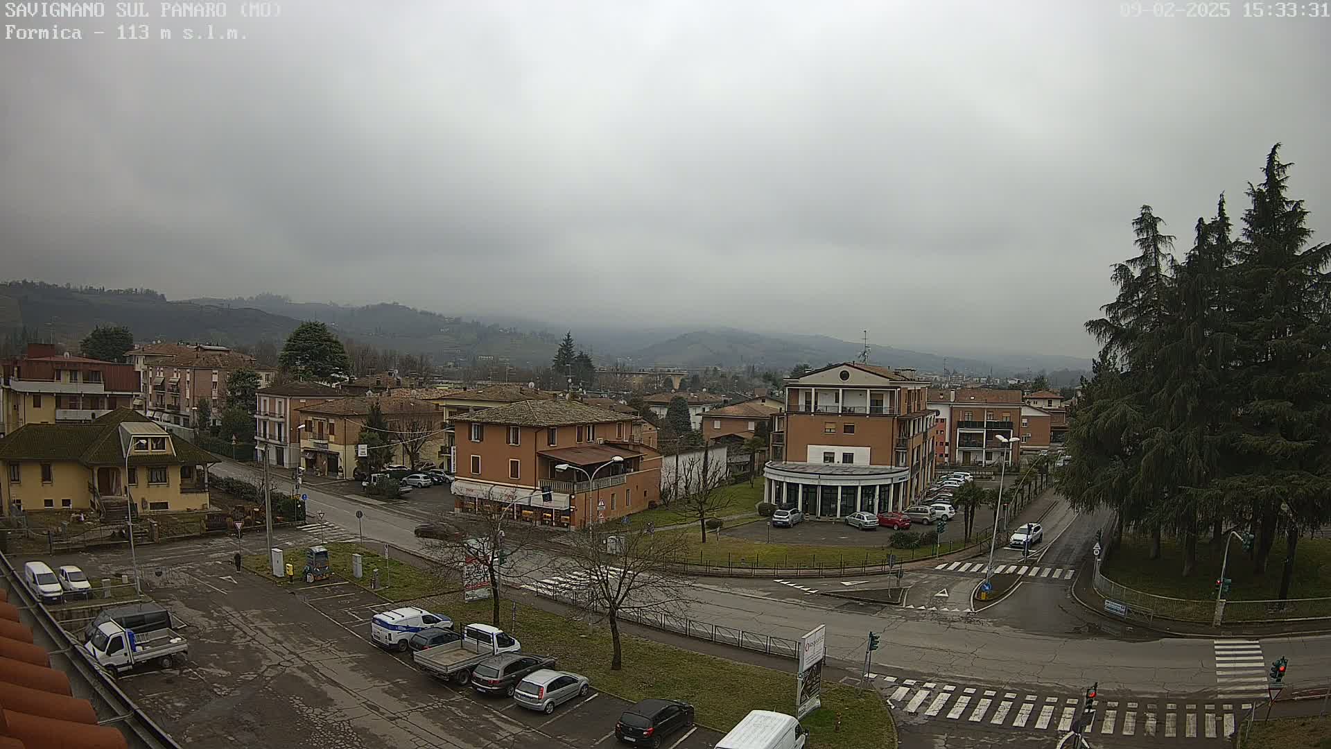 A town with low, light-brown buildings sits in a valley under a cloudy, gray sky; several cars are parked in a lot near a street intersection.