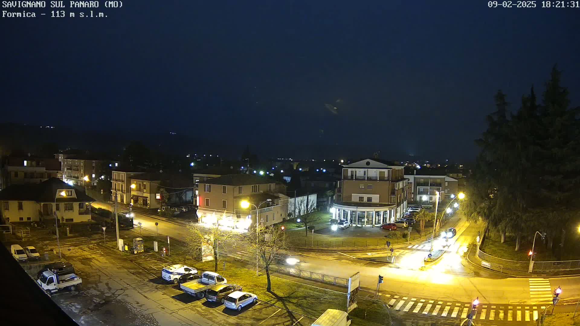 A nighttime aerial view of a town square with several buildings, cars, and streetlights under a dark, overcast sky.