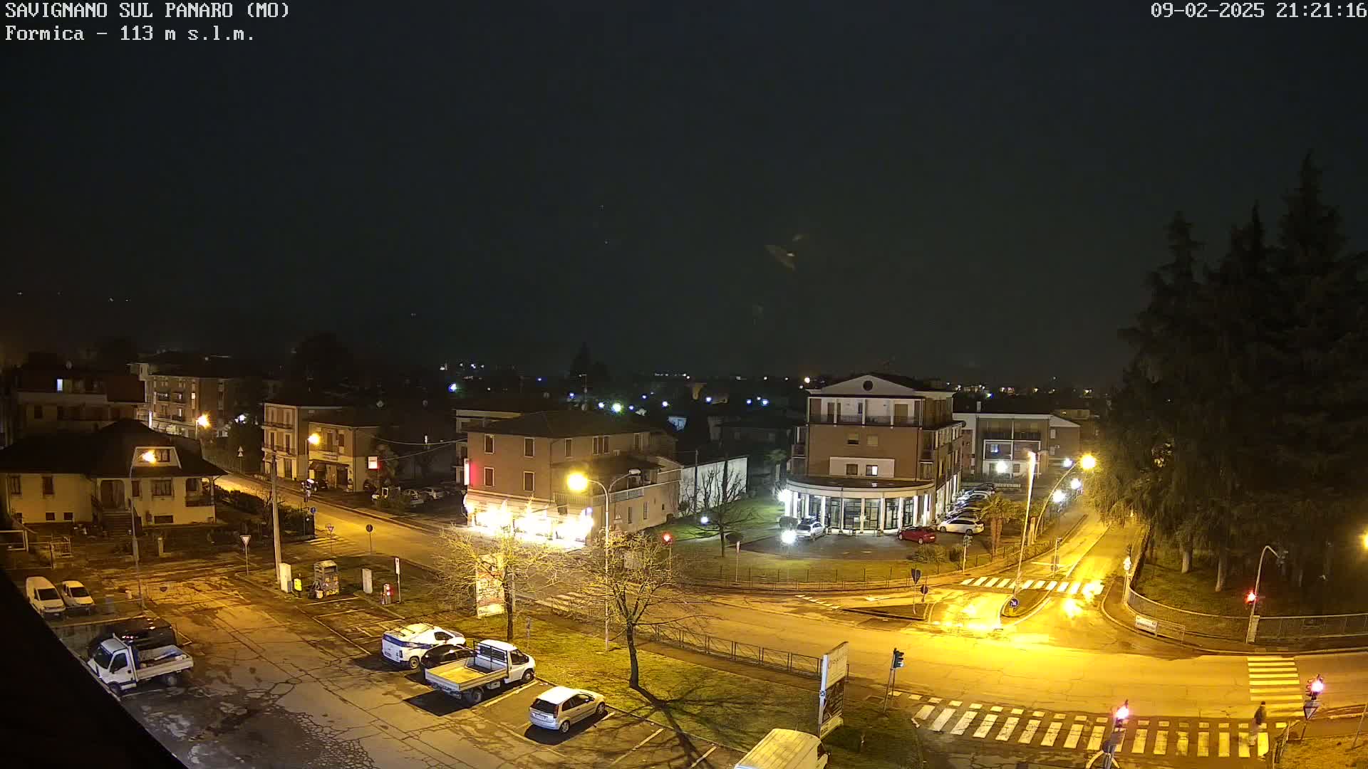 A nighttime, aerial view of a town square with several cars parked and streetlights illuminating the wet streets under a dark sky.