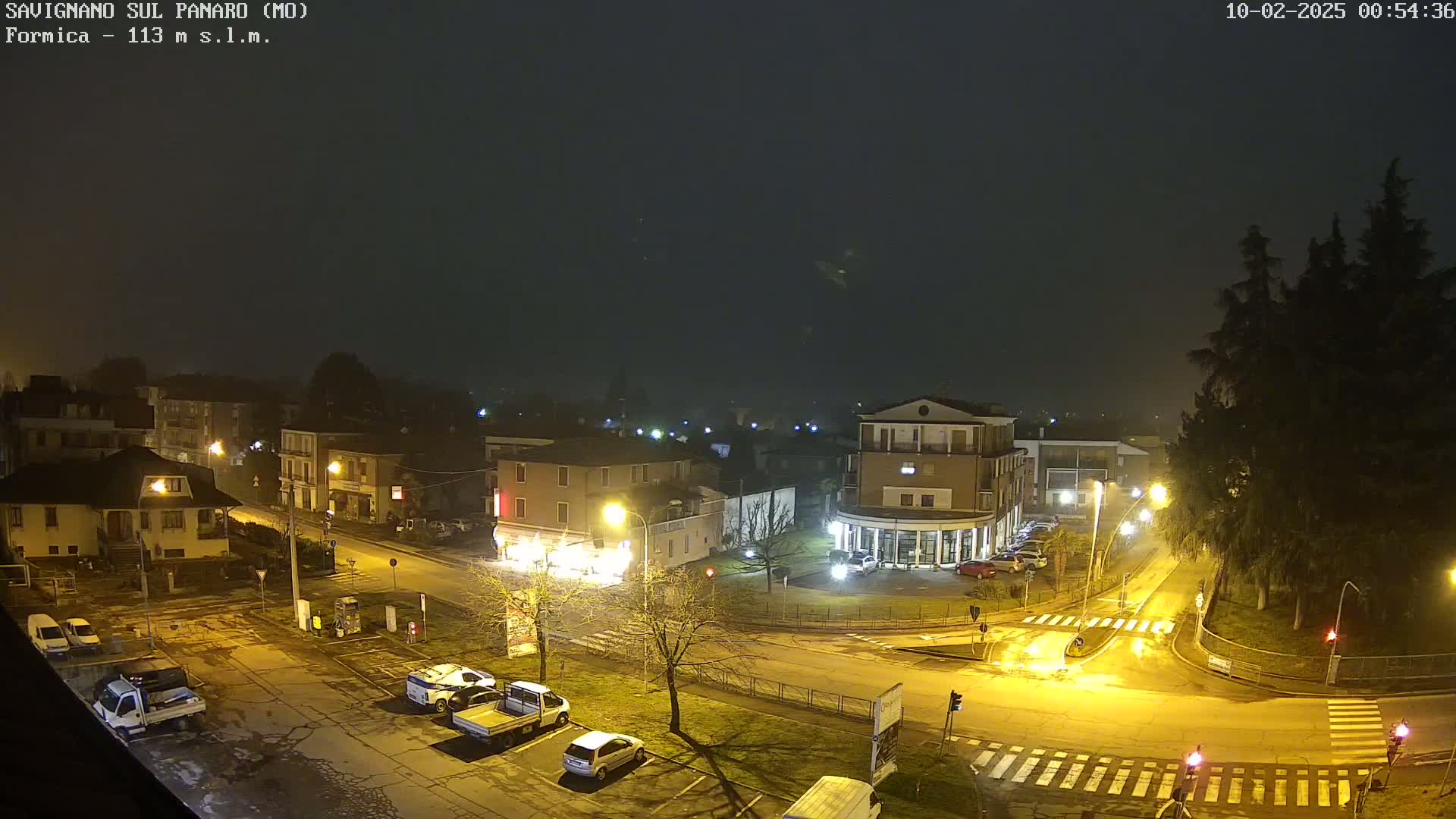 A nighttime, foggy view of a town square with several cars parked and a few buildings lit up.