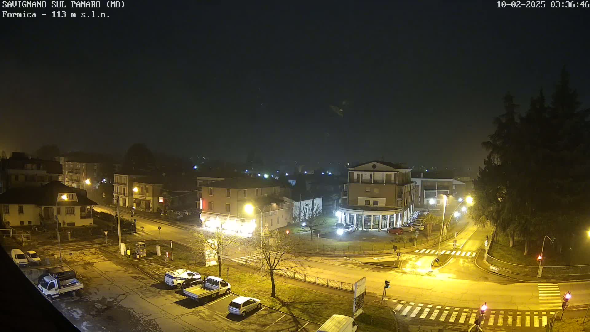 A nighttime, foggy view of a town square with several cars parked, and buildings illuminated by streetlights.