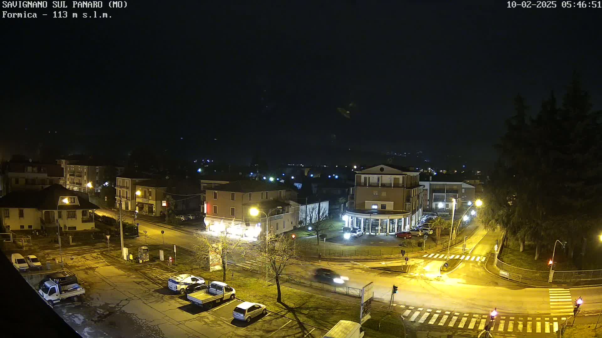 A nighttime aerial view of a town square with several cars parked and driving, under a dark, clear sky.