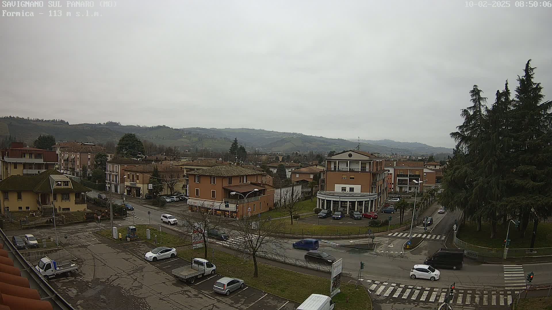A view of a town on a cloudy day, showing several buildings, cars in a parking lot and on the roads, and evergreen trees near a roundabout.