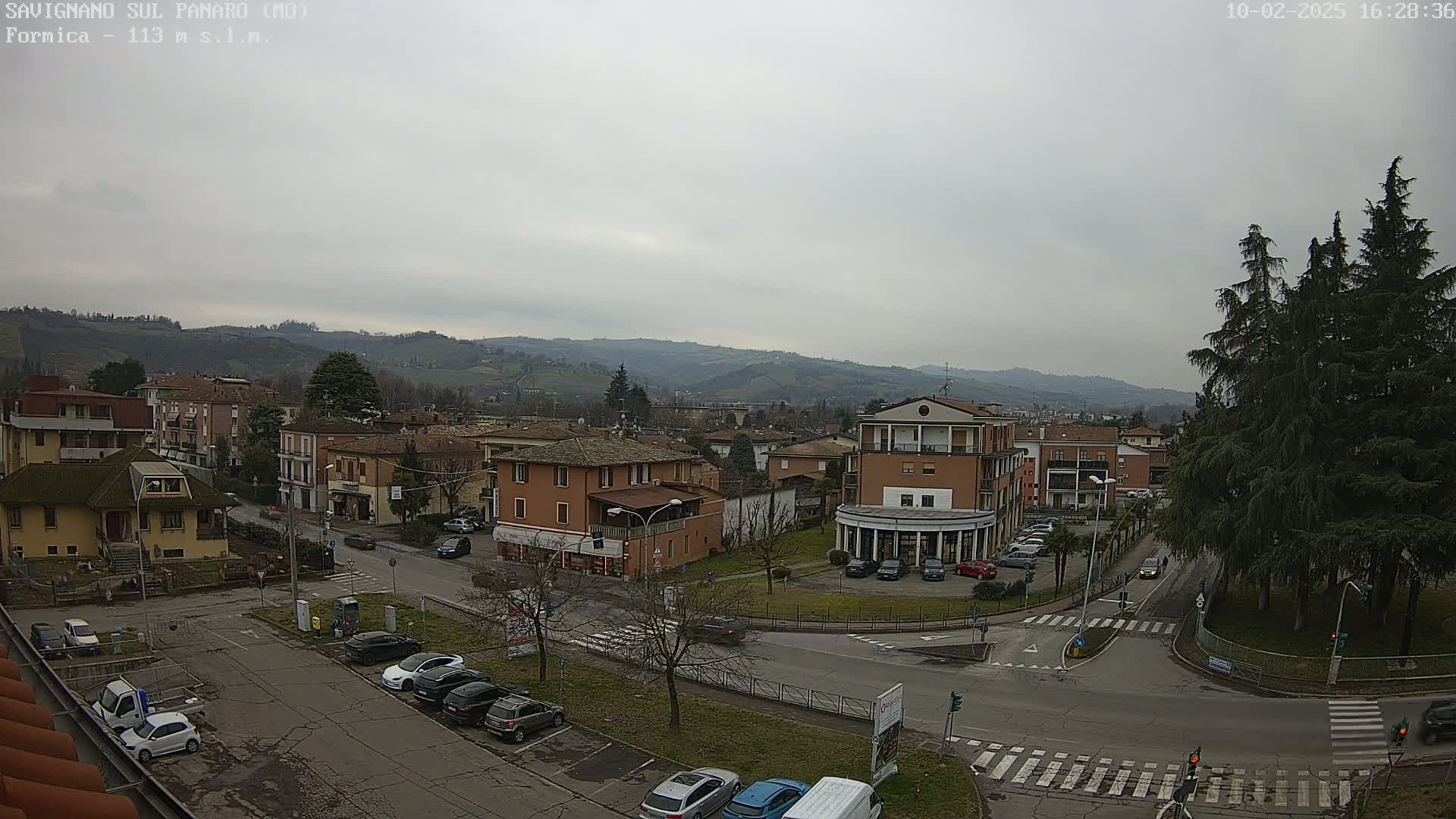 A high-angle, long shot of a town situated in a valley under an overcast sky shows buildings, cars, and evergreen trees.