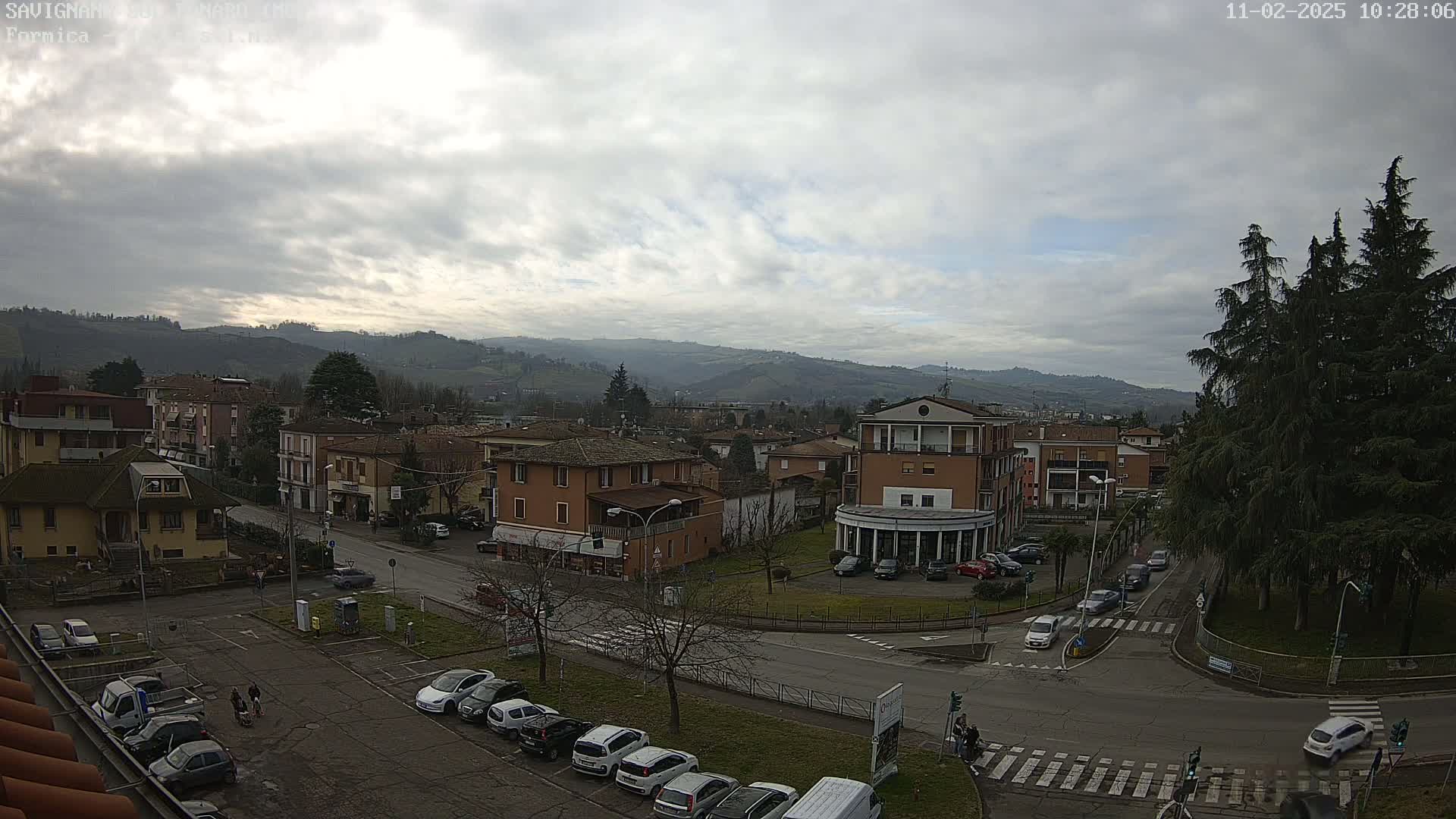 A town nestled in a valley under a cloudy sky, with cars parked in a lot and driving on roads that intersect near a small park.