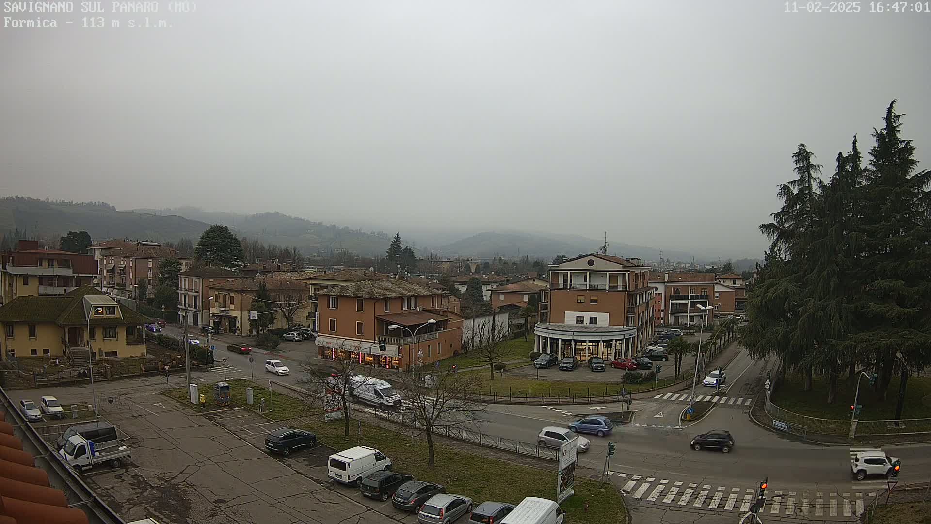 A hazy, overcast day reveals a town square with several buildings, parked cars, and some traffic, all surrounded by hills in the distance.