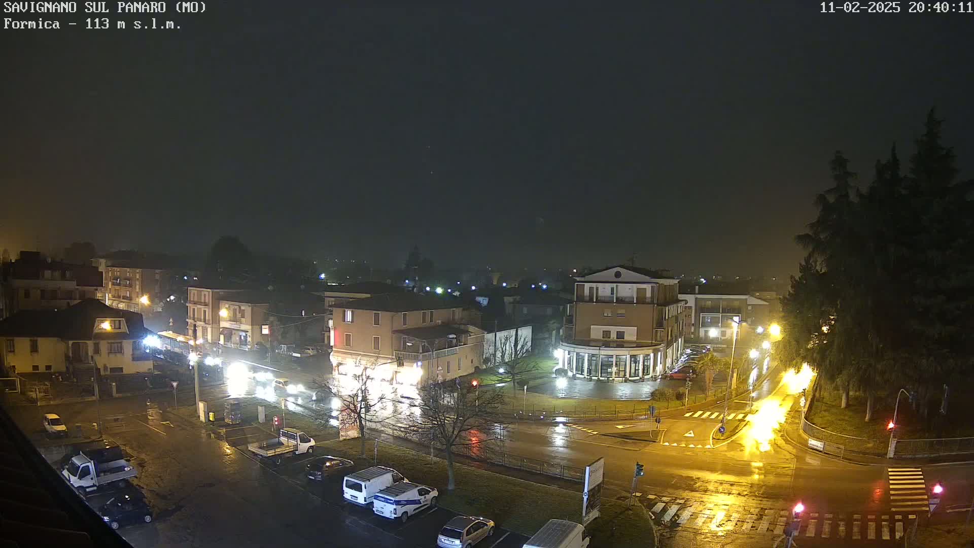 A wet, nighttime view of a town square with several buildings, vehicles, and streetlights illuminated under a dark, overcast sky.