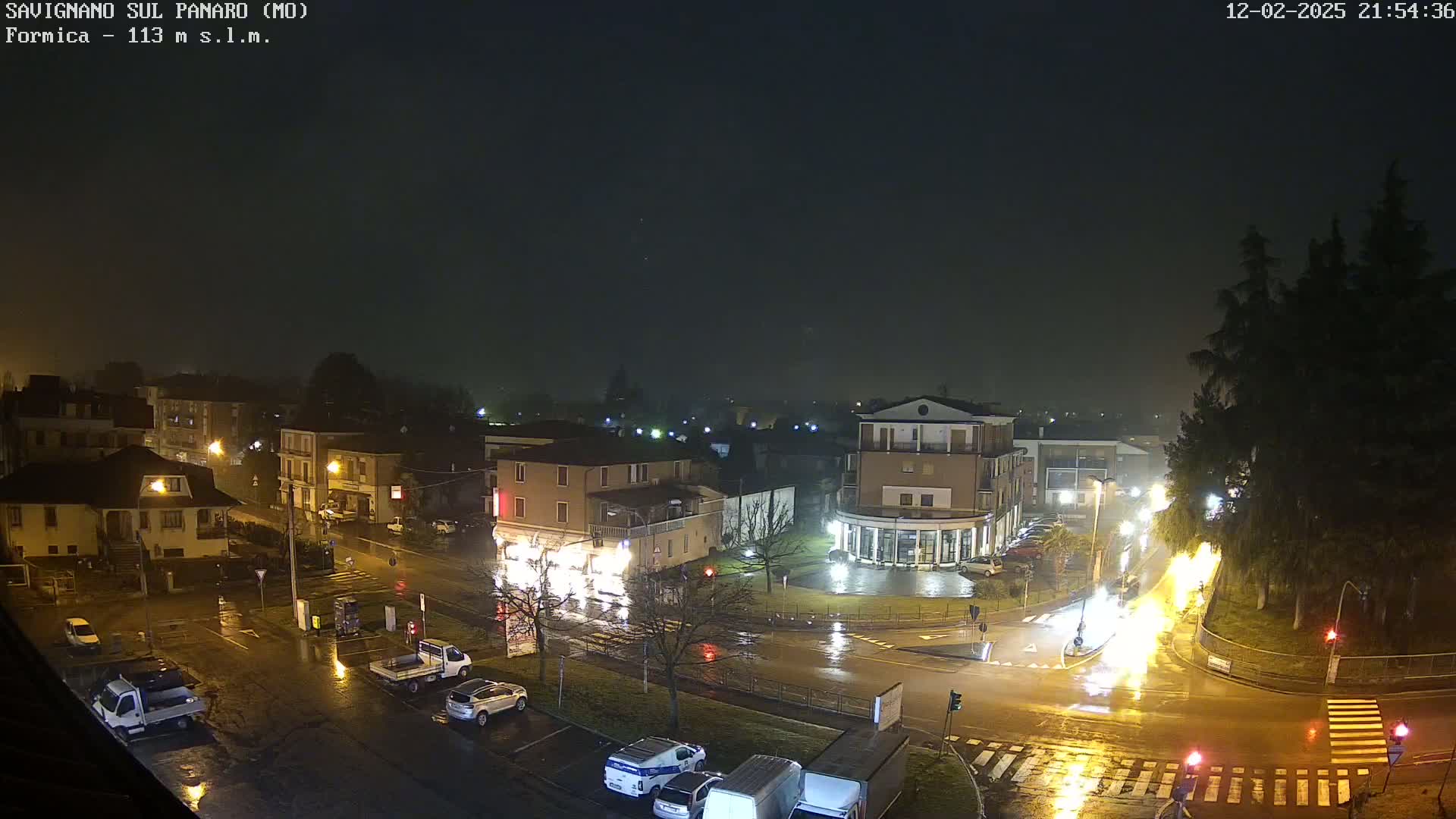 A rain-slicked town square at night is illuminated by streetlights, with several cars parked and driving on wet roads.