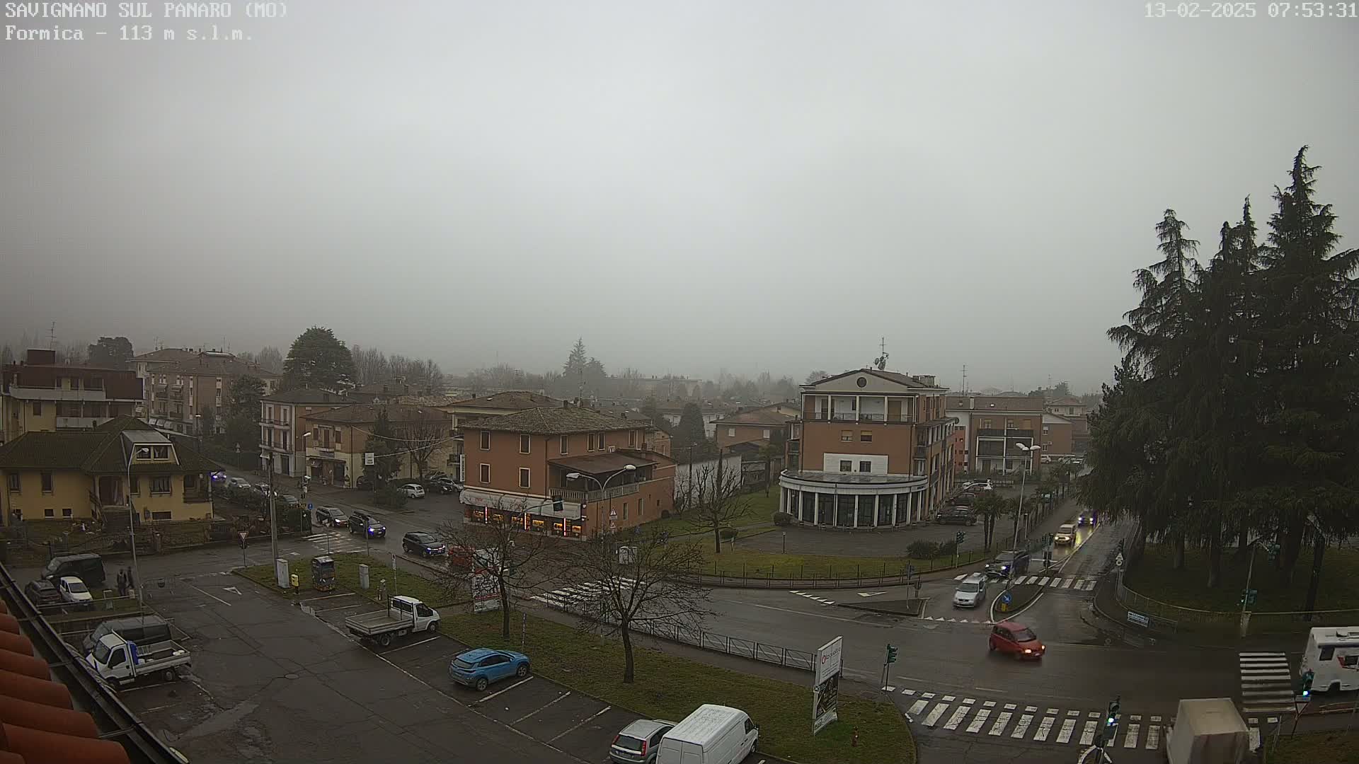 A foggy, overcast day reveals a view of a town square with several buildings and cars, surrounded by trees and other vegetation.