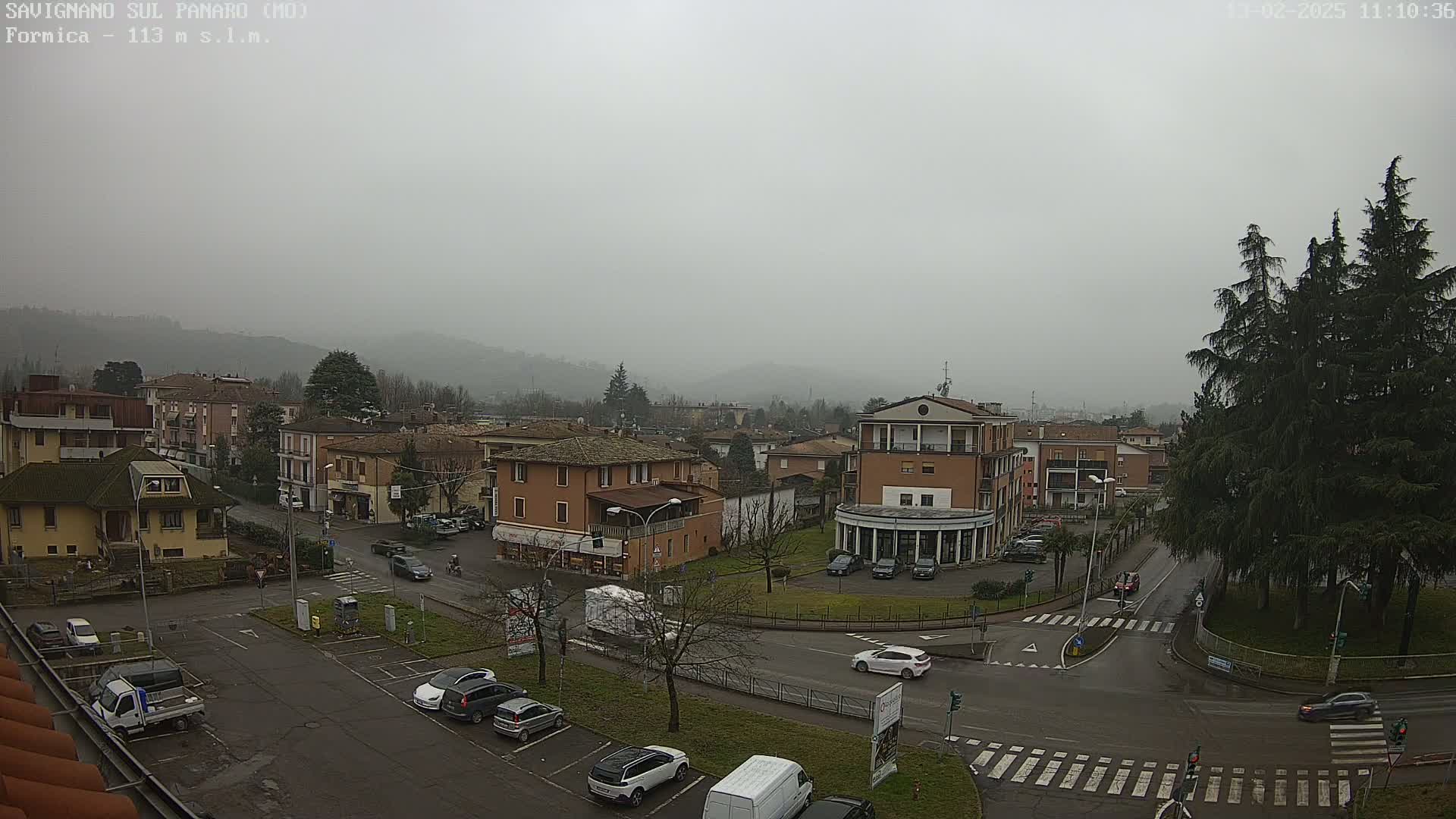 A hazy, overcast day reveals a town square with cars parked and driving around it, surrounded by low buildings, and a few evergreen trees in the foreground.